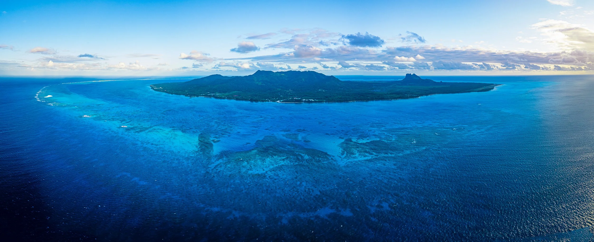 Aerial panorama view of an island with mountains, green land, and surrounding blue ocean with coral reefs and shallow waters near the coast