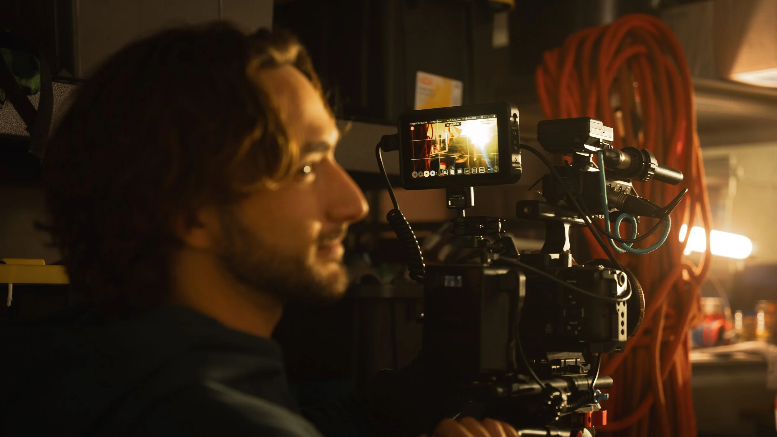 A person operating a professional video camera setup in a dimly lit environment with a warm light source in the background.