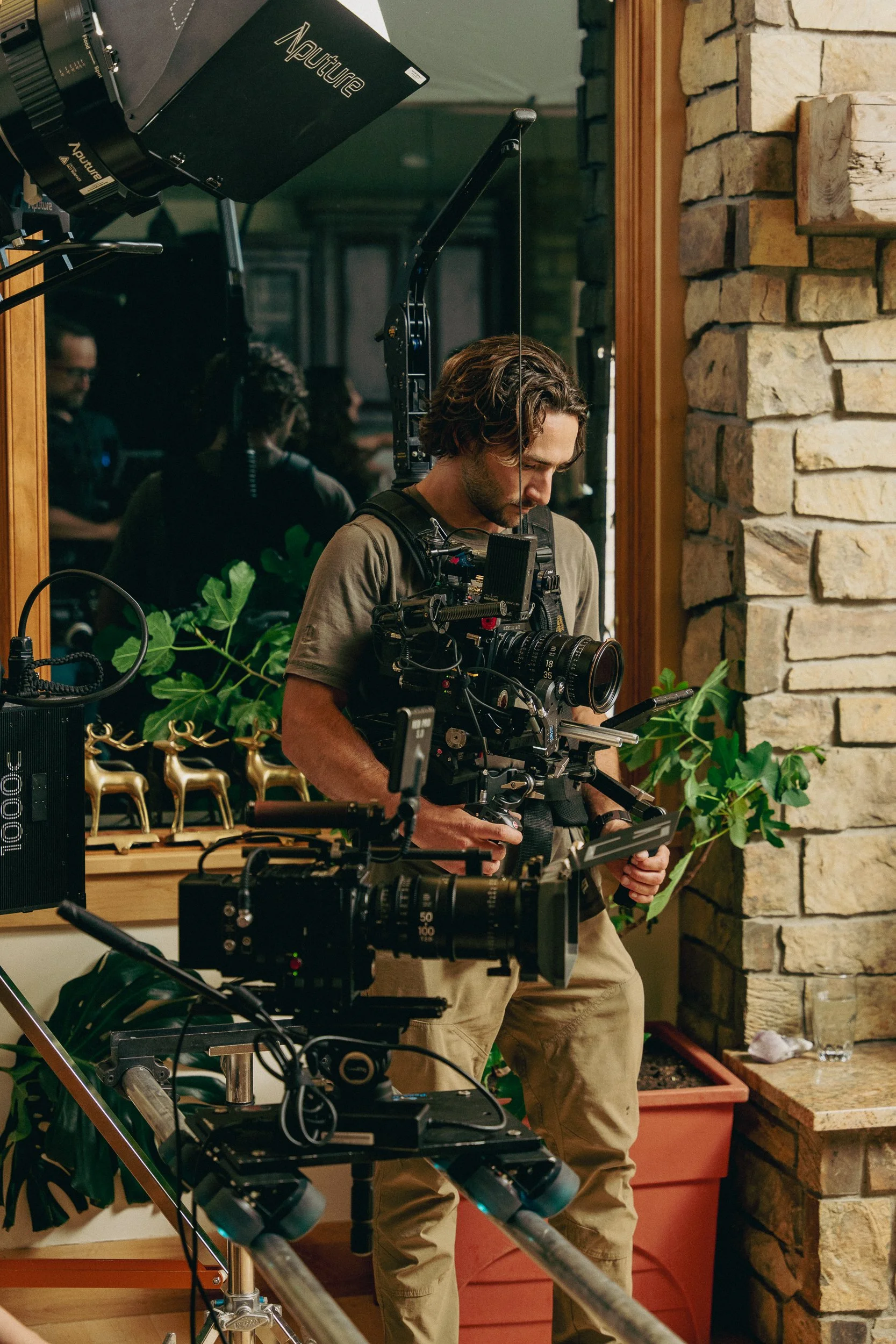 A videographer dressed in beige pants and a green shirt is operating a professional camera (RED 8 K Helium Camera) with stabilizer (Steadicam) equipment indoors. There are film lights, a camera slider with another RED Scarlet on it.
