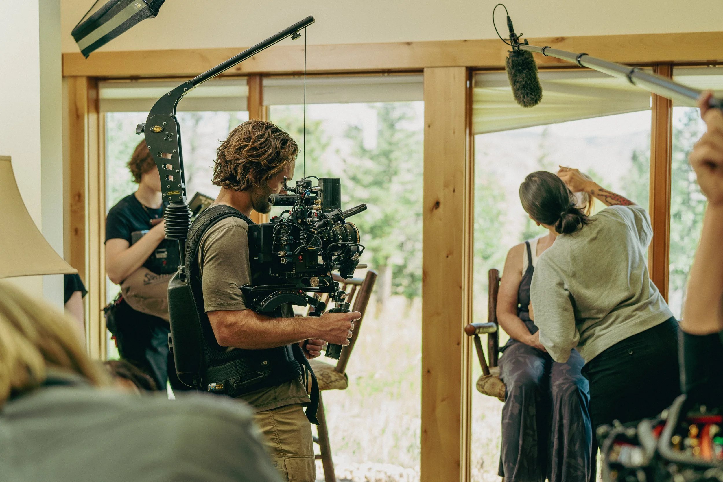 Film crew recording a scene indoors with a woman sitting by a window, a person with a camera, and crew members with equipment, during daylight.