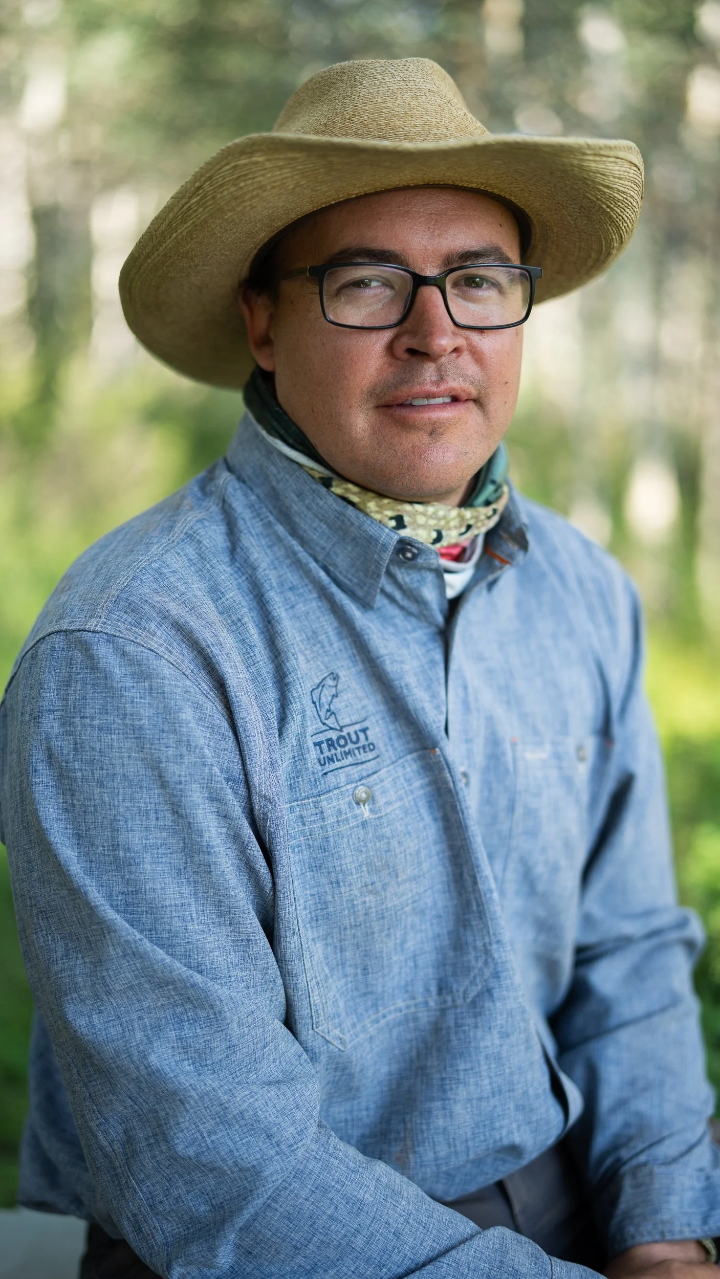 Man wearing a wide-brimmed straw hat, glasses, a blue long-sleeve shirt with a "Trout Unlimited" logo, and a neck gaiter, outdoors in a wooded area.