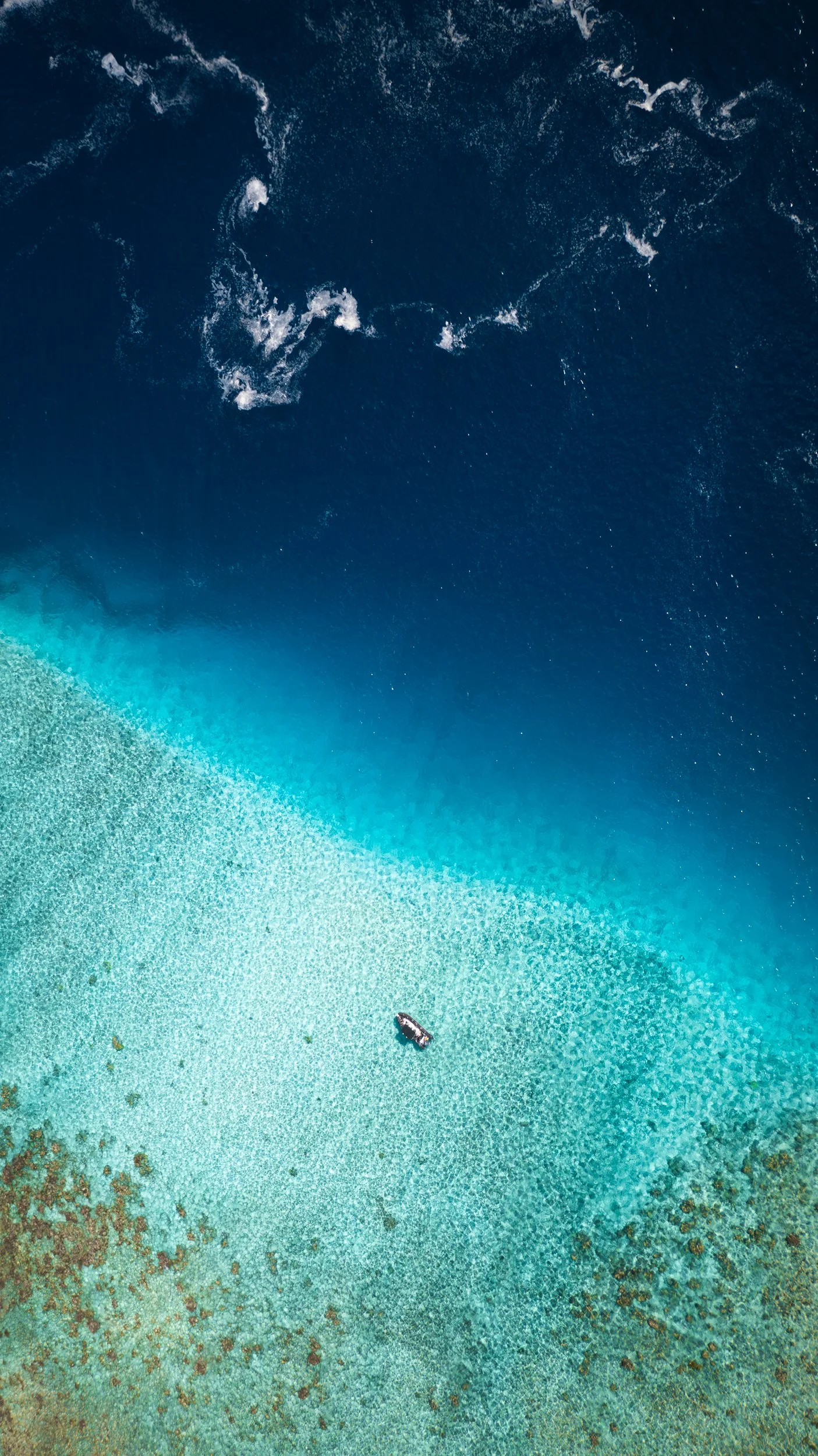 A professional Aerial drone image of a Marine Biology NGO research boat anchored in a turquoise coral reef shallow area near the ocean, with deep blue water and some waves in the background. Ocean Conservation non-profit, NGO