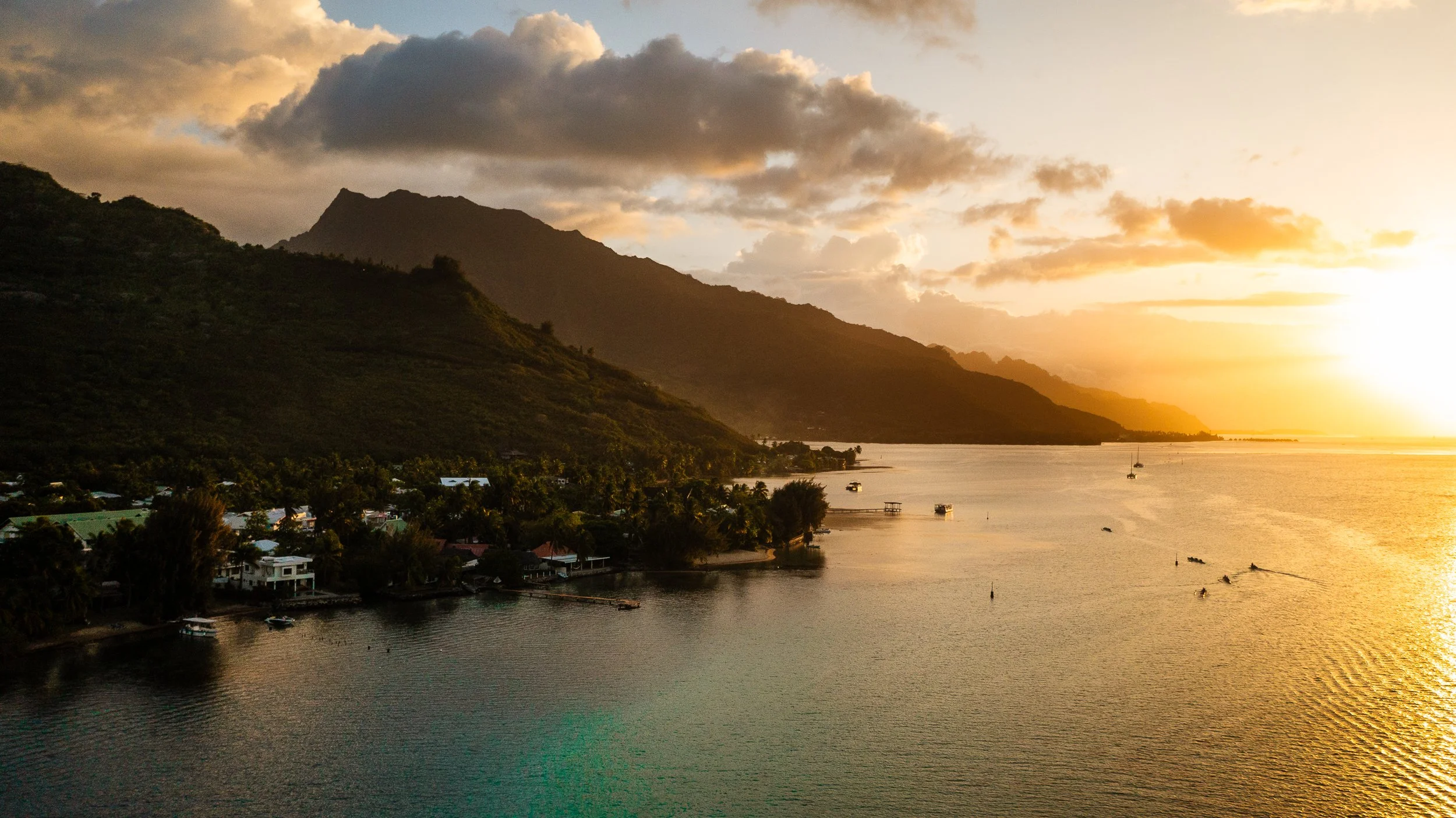 A professional Aerial drone image of a Sunset over a bay with mountains in the background, featuring houses along the shore and boats in the water.