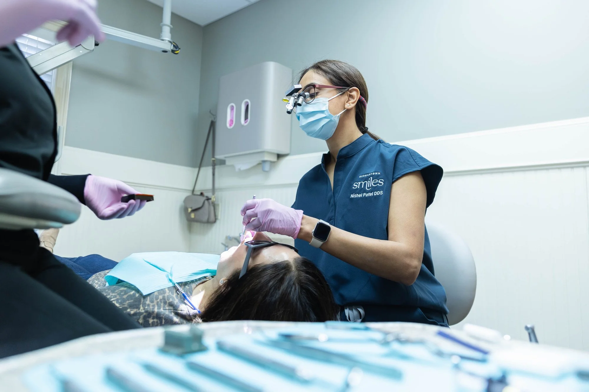 Dentist performing a dental procedure on a patient lying in a dental chair, with dental tools on a tray in the foreground.