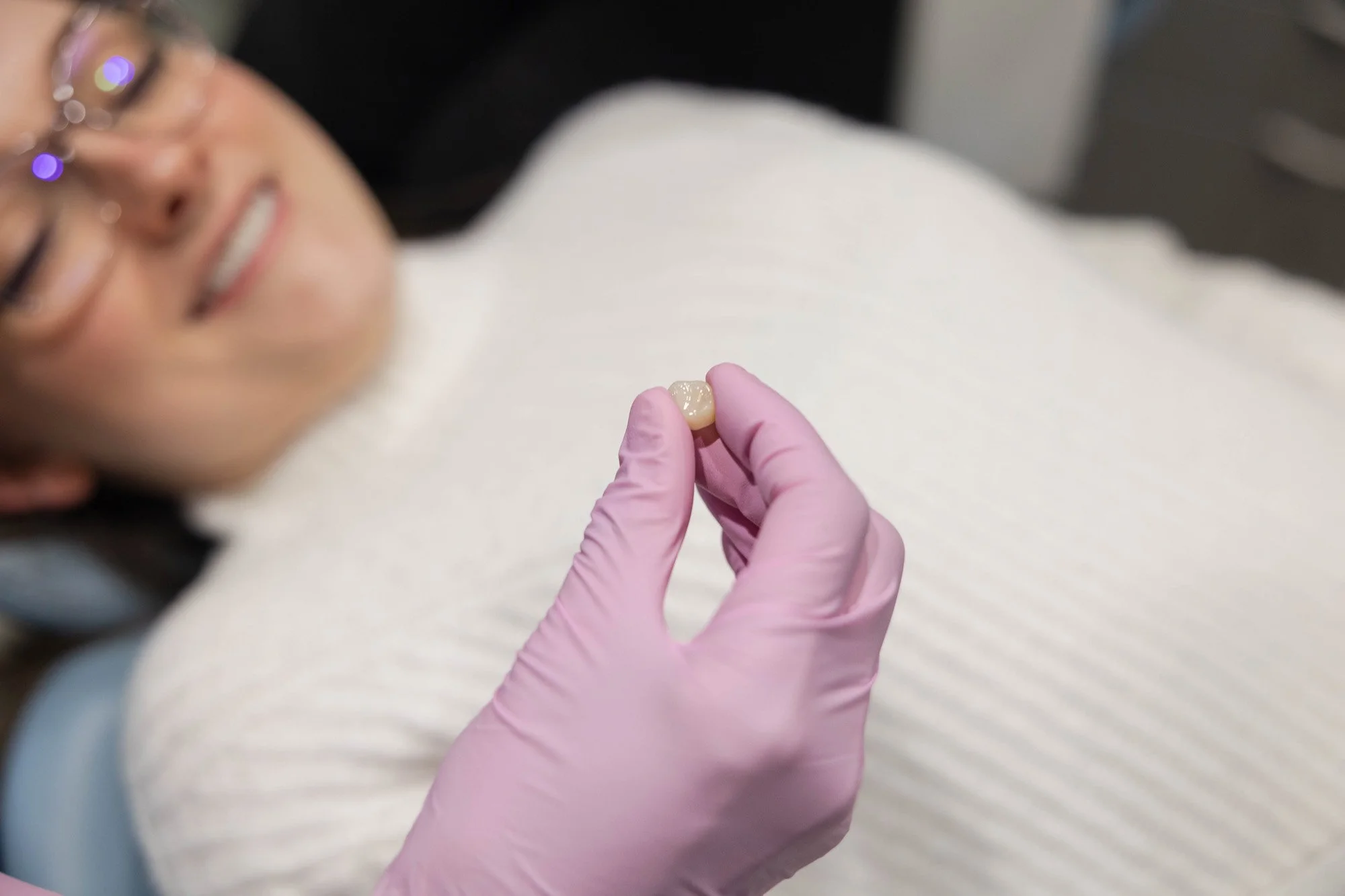A dentist wearing pink gloves holding a small dental crown or cap near a patient's mouth, who is smiling and lying back in the dental chair.
