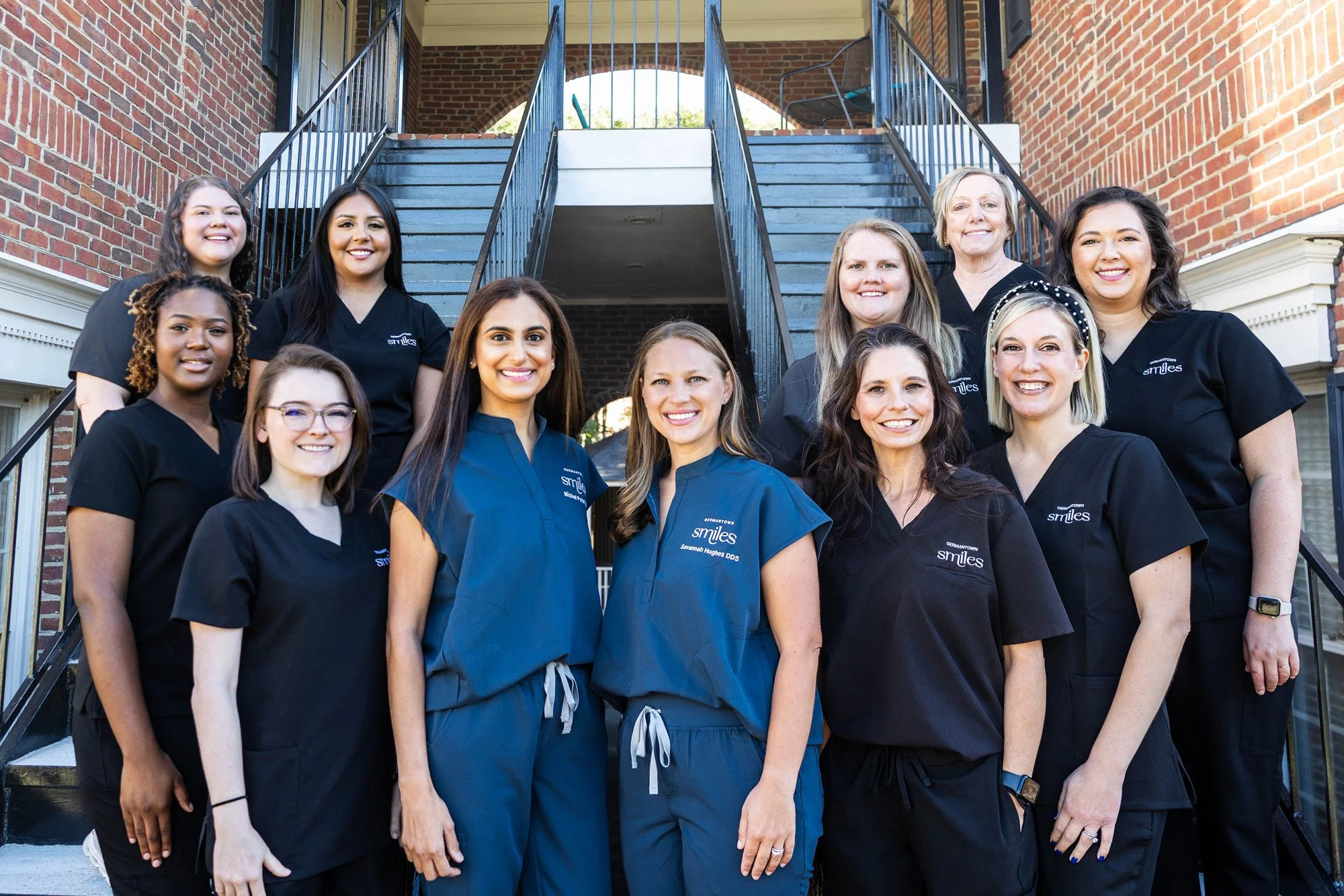 Group of dental professionals standing on outdoor staircase in front of brick building, wearing scrubs, smiling at camera.
