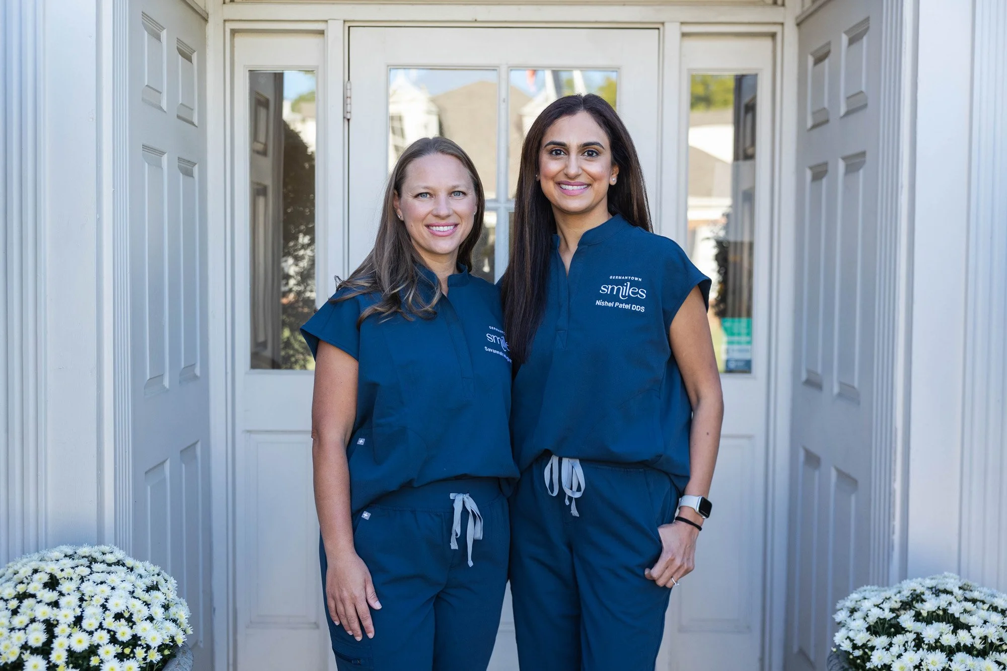 Two women wearing navy medical scrubs standing outside a door with glass panels, smiling at the camera, with white flower arrangements on either side.