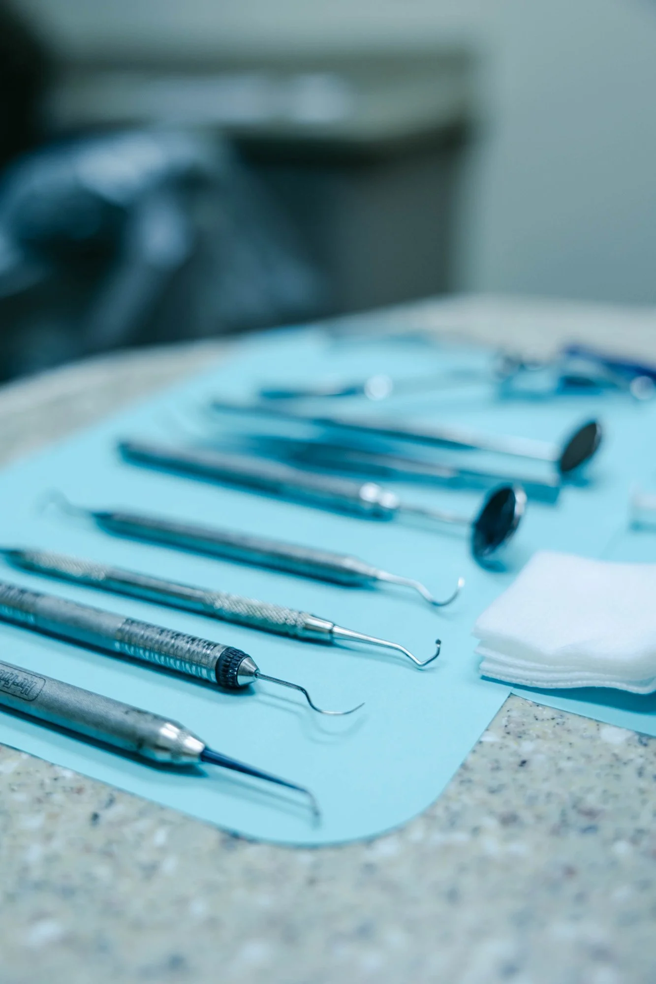 Dental tools arranged on a blue tray, including picks, mirrors, and possibly a syringe tip, with a stack of gauze and a blurred background.