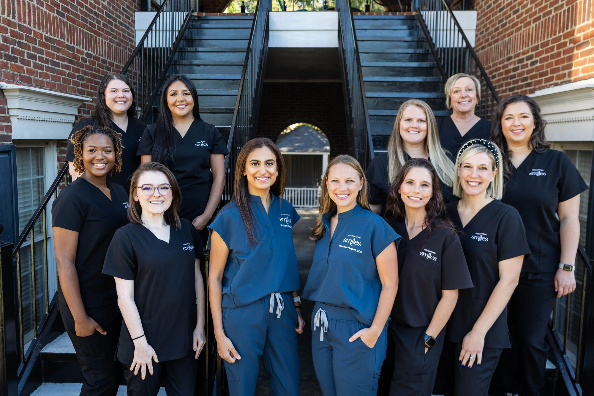 Group photo of Smiles dental team members standing outdoors on stairs in front of brick building, wearing black and blue scrubs.