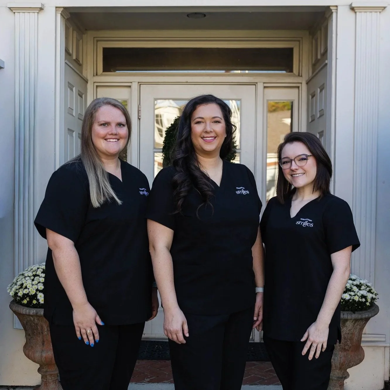 Three women in black uniforms standing outside in front of a house with white siding and a door. They are smiling and looking at the camera.