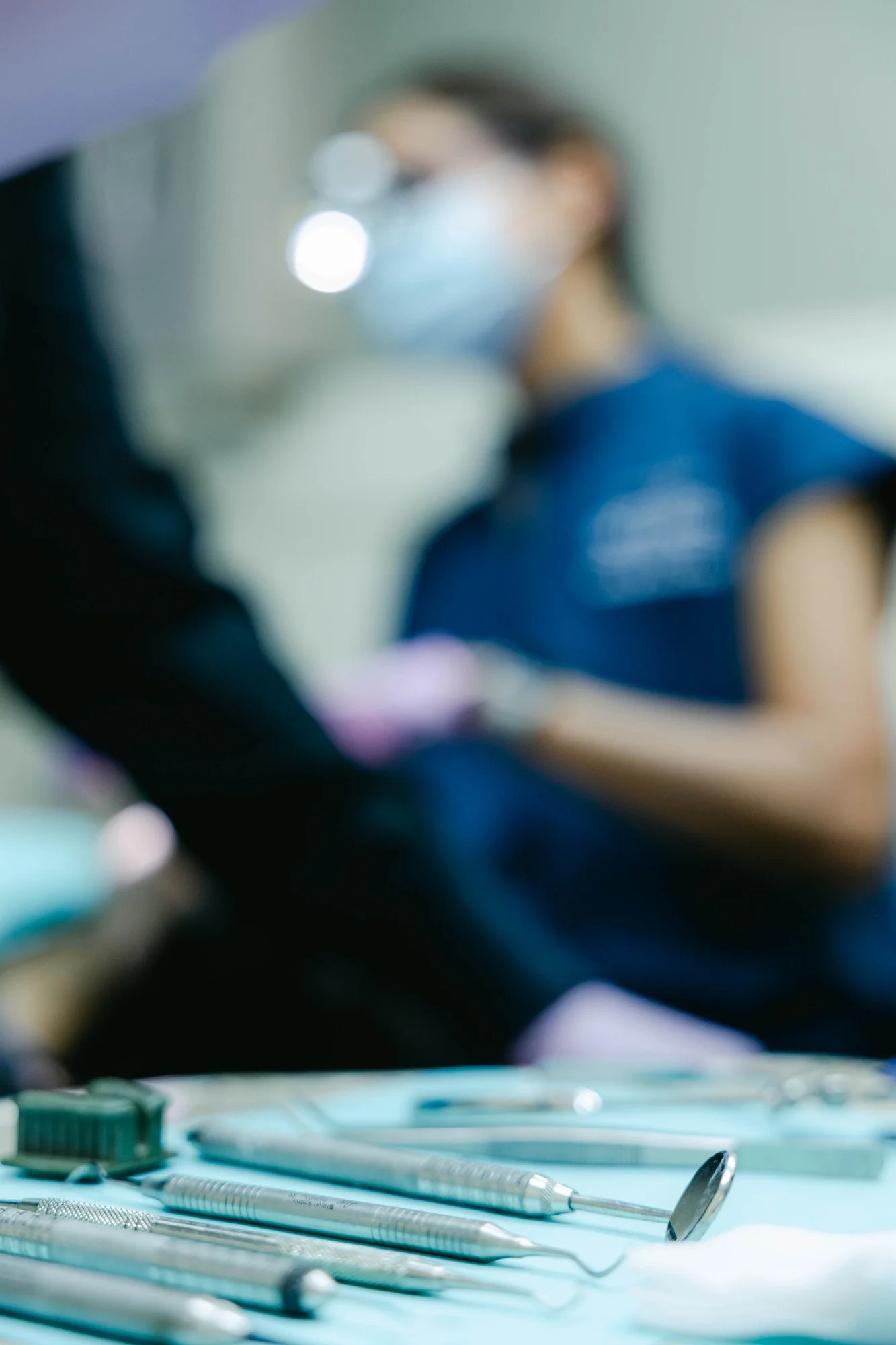 A blurred person wearing a face mask and glasses in a medical setting, with dental tools in the foreground.