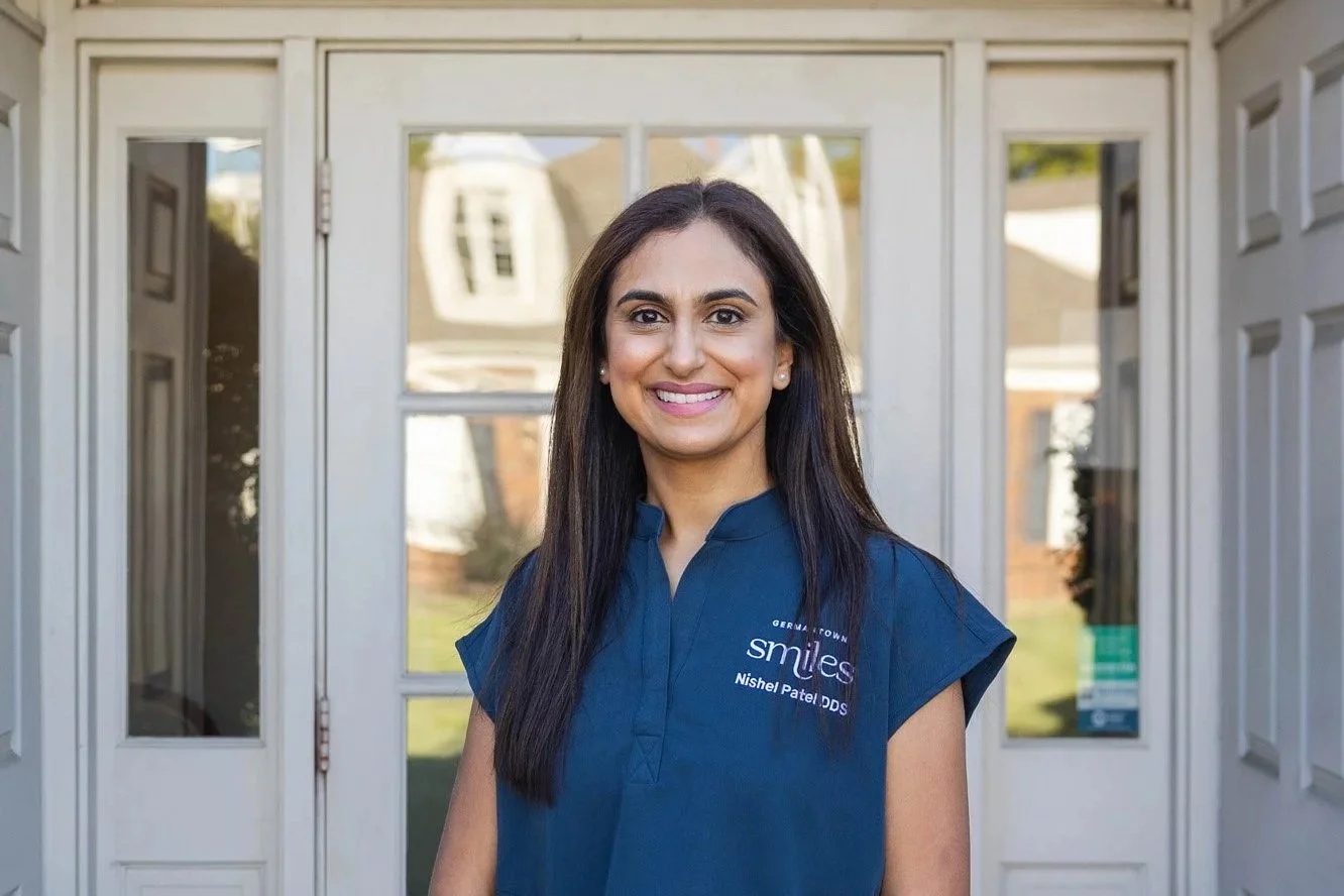 A woman with long dark hair smiling in front of a doorway, wearing a blue shirt with 'smiles' and 'Nishel Patel DDS' embroidered on it.
