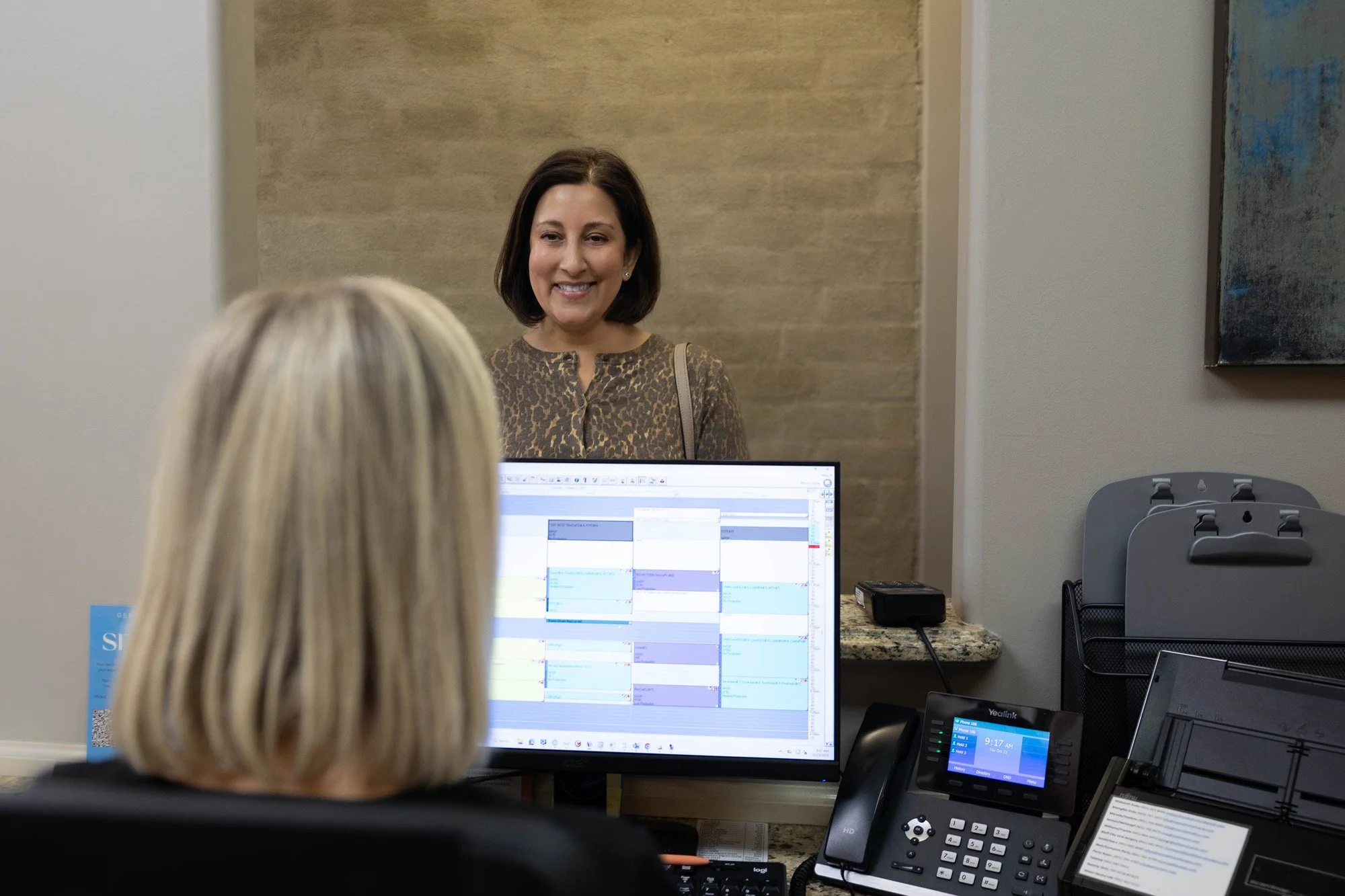 A woman smiling at a desk in an office, seen through a half wall, with another woman sitting at the desk facing a computer monitor.