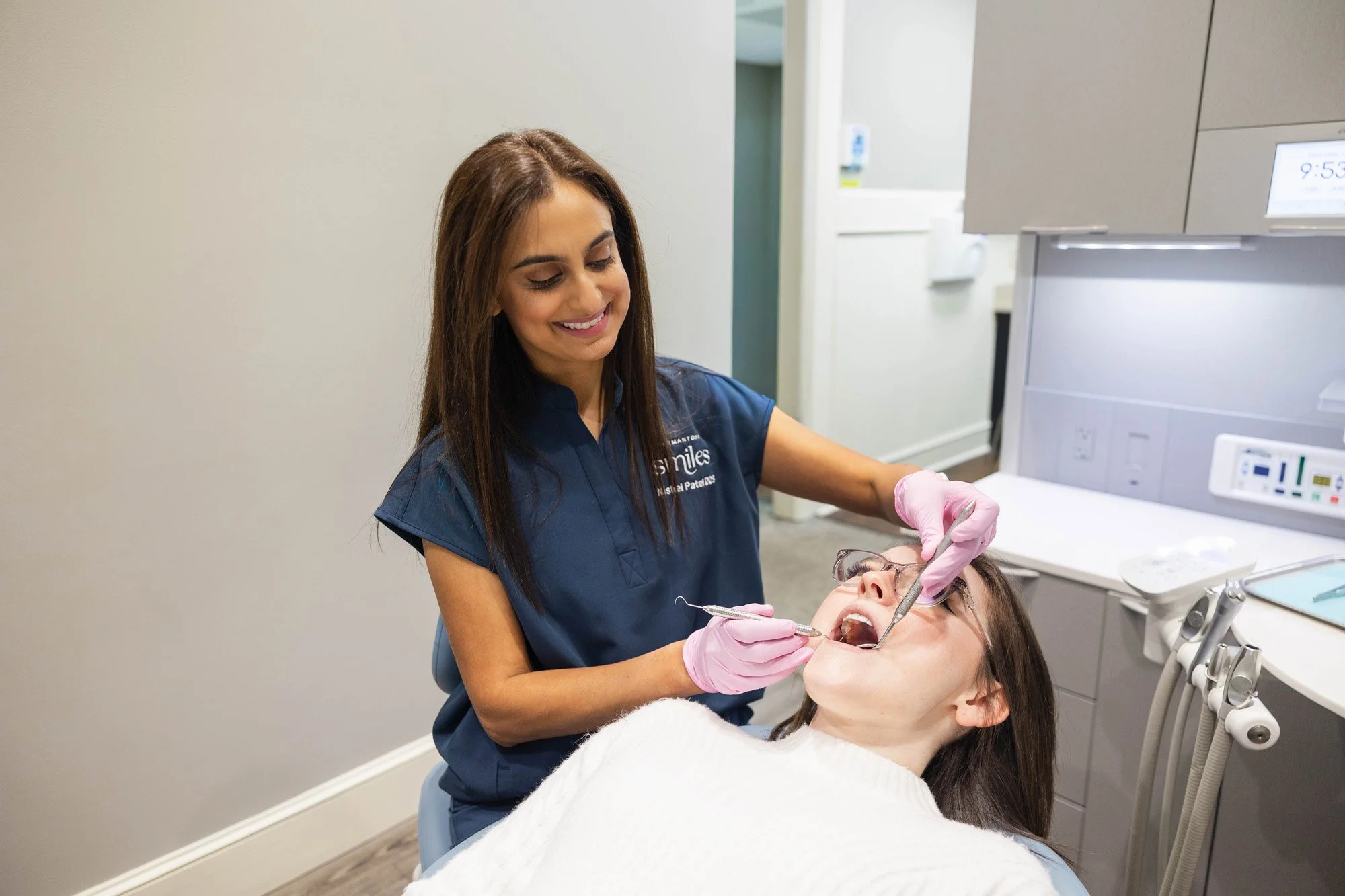 A female dentist in blue scrubs and pink gloves examining a woman's teeth in a dental office.