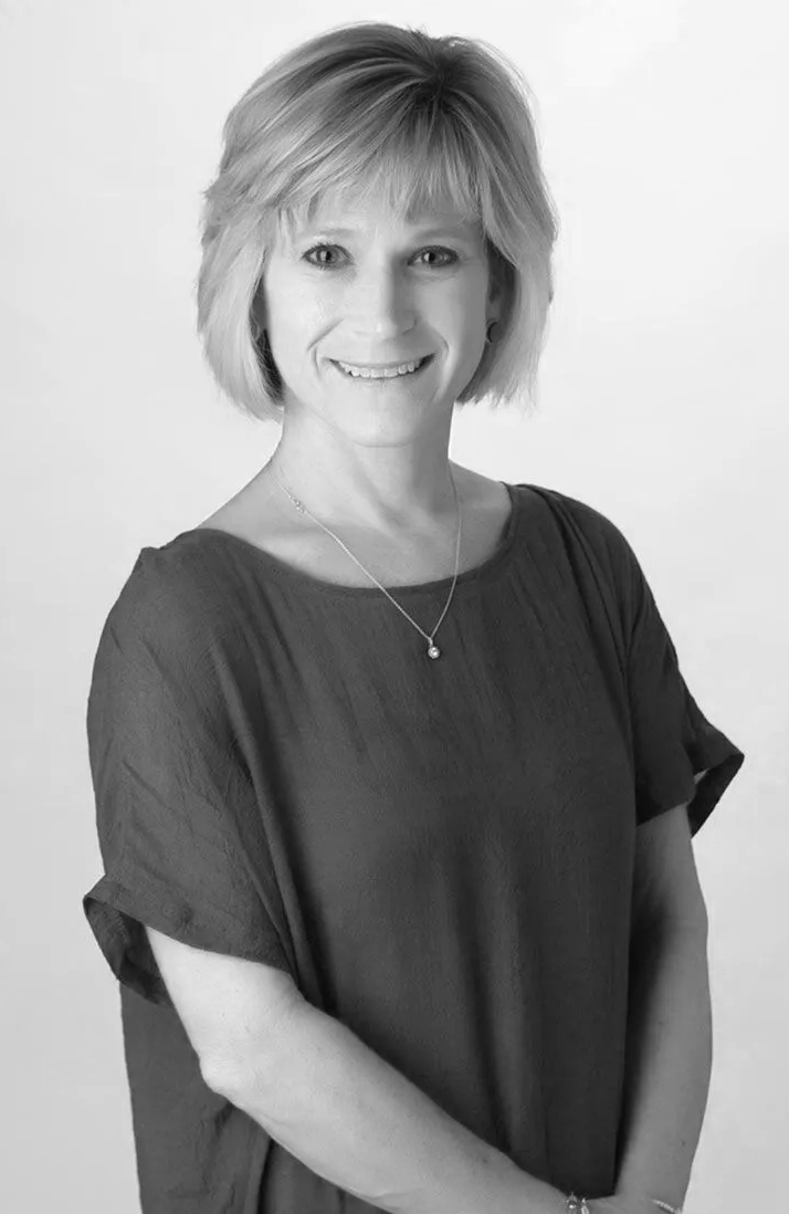 A smiling woman with short, layered hair, wearing a dark blouse, a necklace with a small pendant, and a bracelet, standing against a plain background.