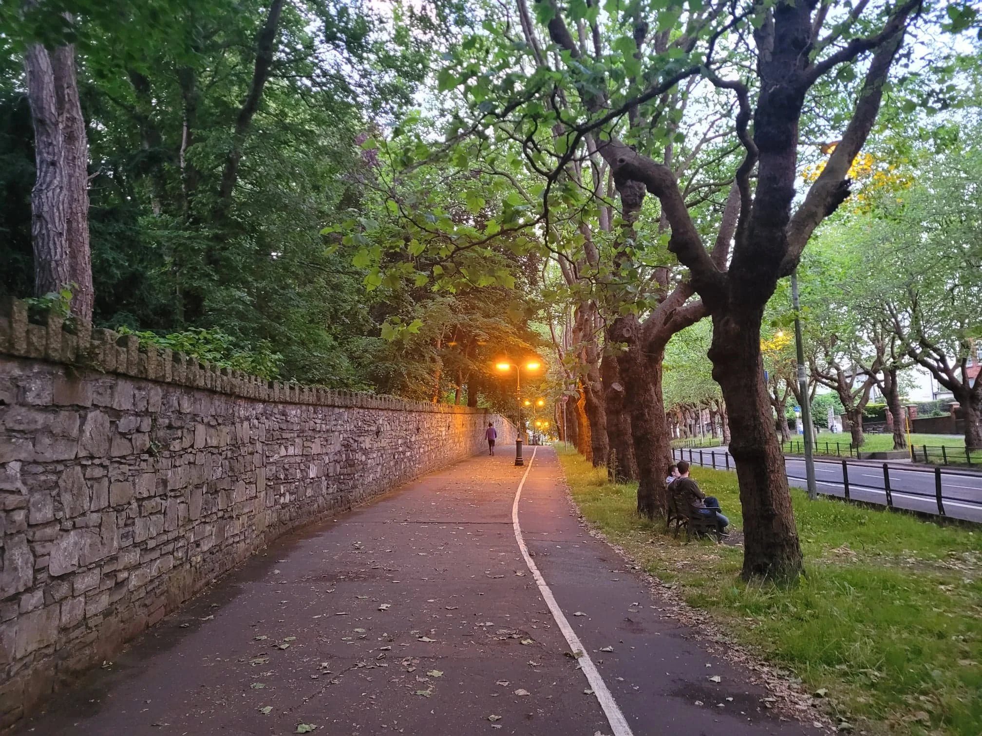 A peaceful park pathway lined with trees on one side and a stone wall on the other, with street lamps illuminating the area at dusk. A person is sitting on a bench, and another person is walking down the sidewalk.
