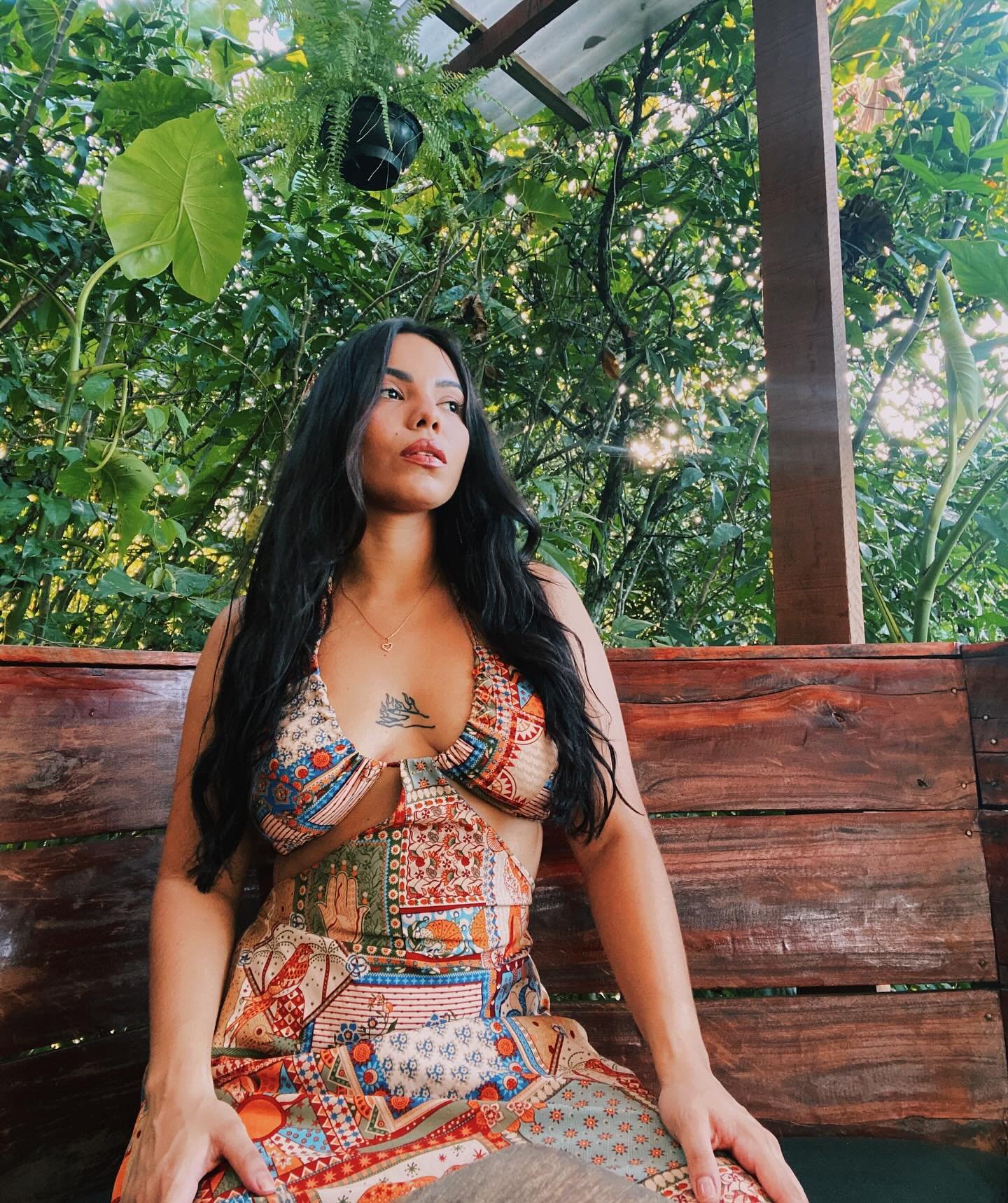 Mulher de cabelo comprido e escuro, usando vestido colorido com detalhes em crochê, sentado em banco de madeira, com vegetação densa ao fundo e iluminação natural.