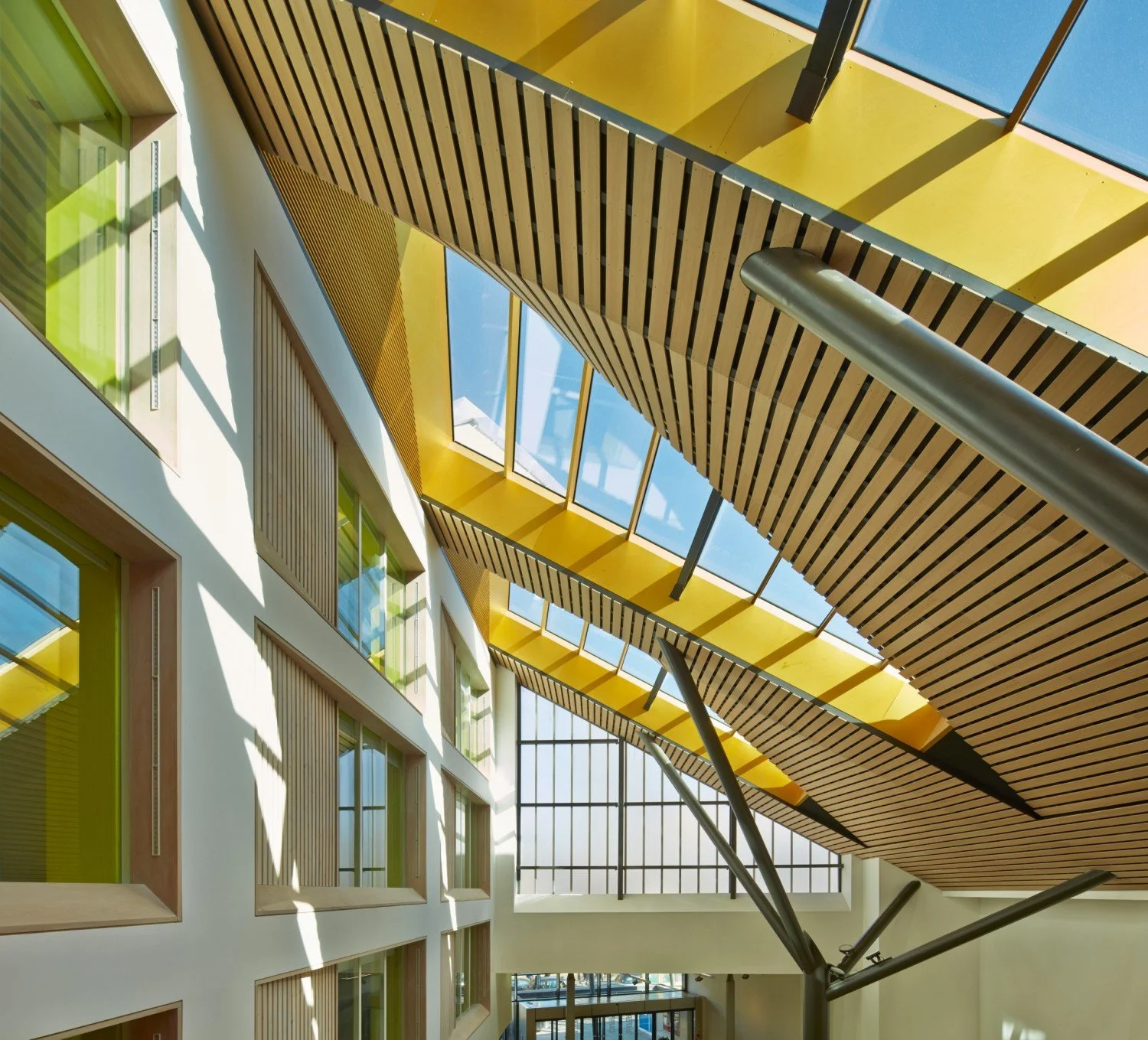 Interior view of a modern building with a high ceiling featuring wooden slats and large windows with yellow and green frames, sunlight casting shadows.