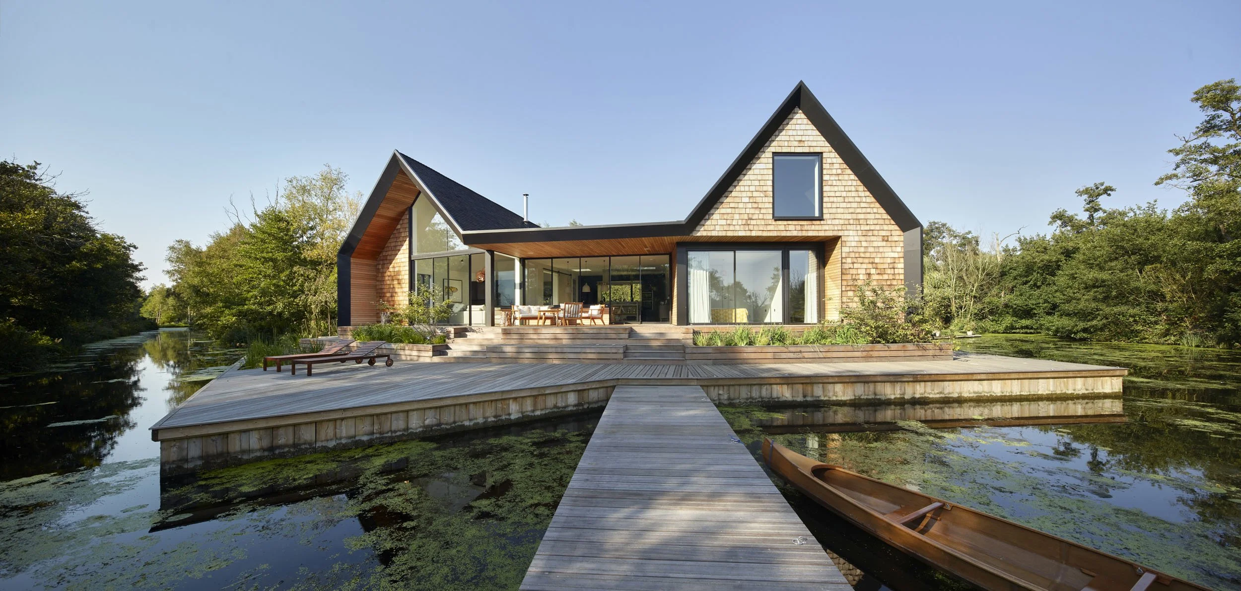Modern house with large glass windows and a wooden exterior built on stilts over a pond with a dock and a canoe, surrounded by trees.