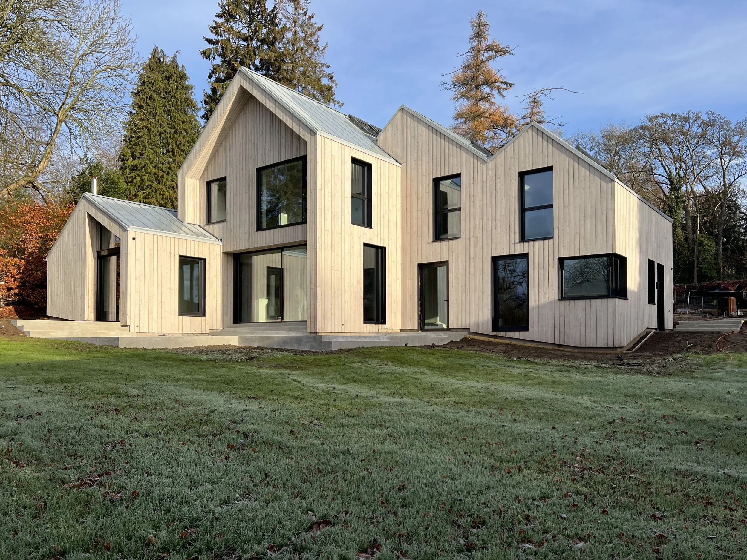 A modern house with a wooden exterior, large windows, and a metal roof, surrounded by trees and a grassy yard.