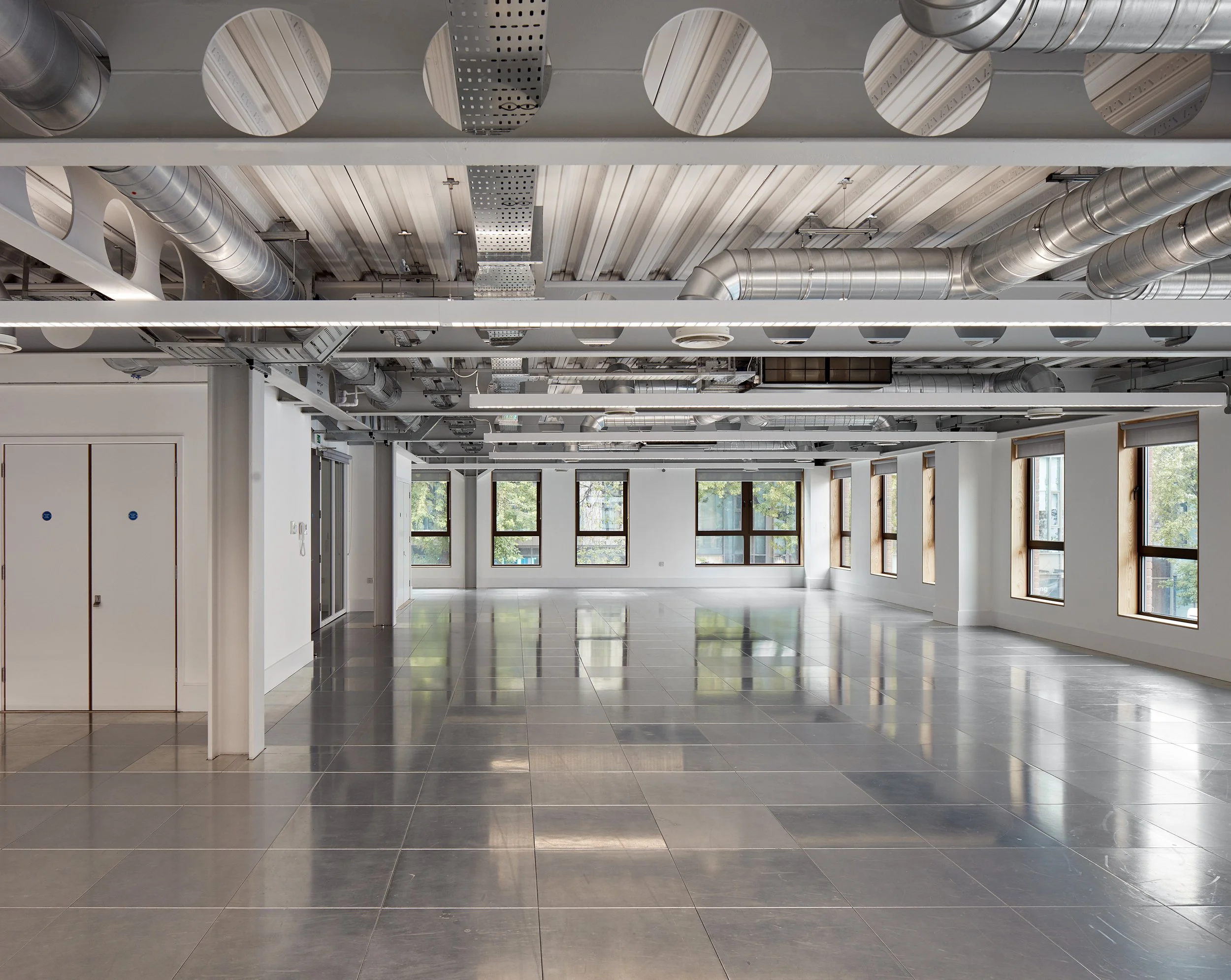 Empty modern office space with large windows, gray tiled flooring, white walls, visible ceiling with exposed silver ductwork and drop ceiling panels, bright natural light.