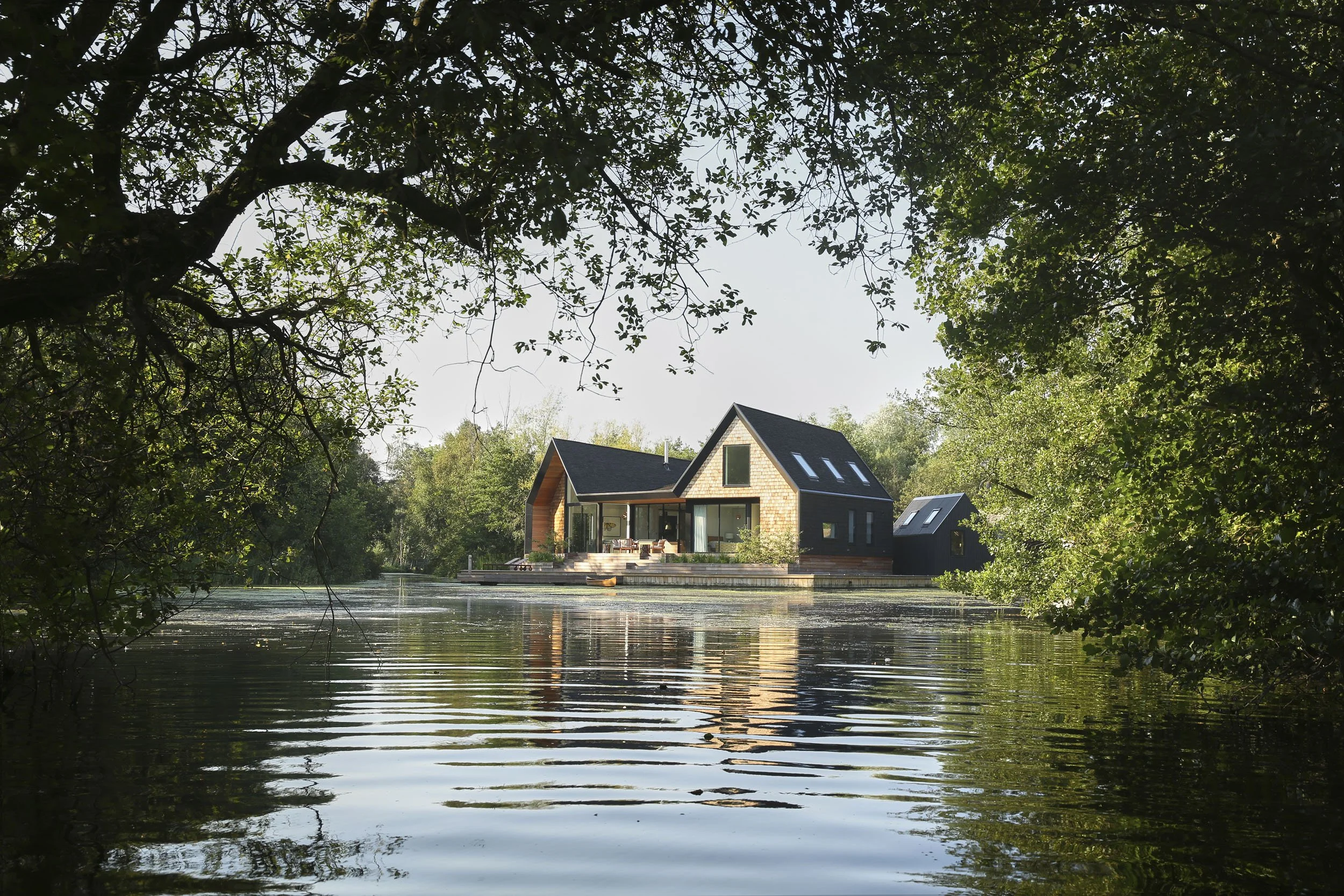 Modern house with large windows and a deck by a calm lake, surrounded by trees with dense foliage, reflecting on the water.