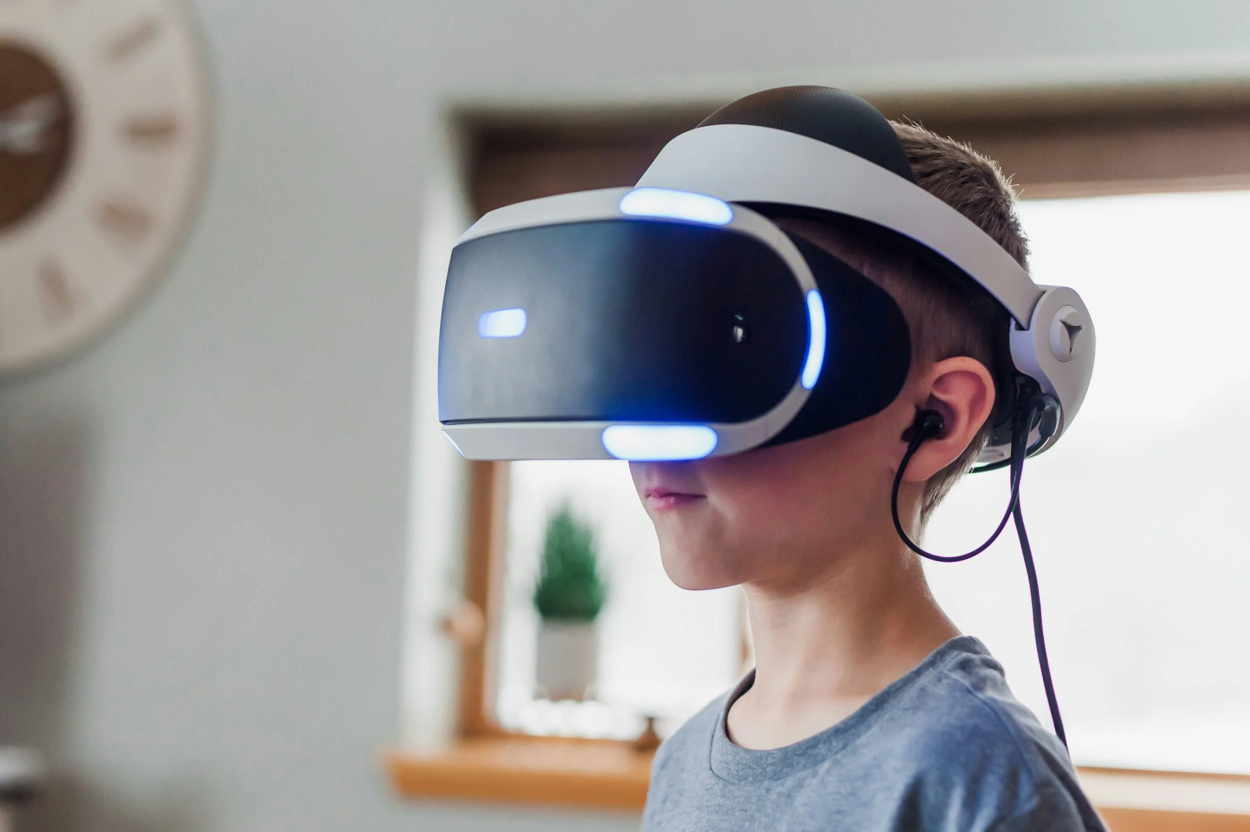 Young boy wearing a gray shirt using a virtual reality headset with blue lights, standing indoors.