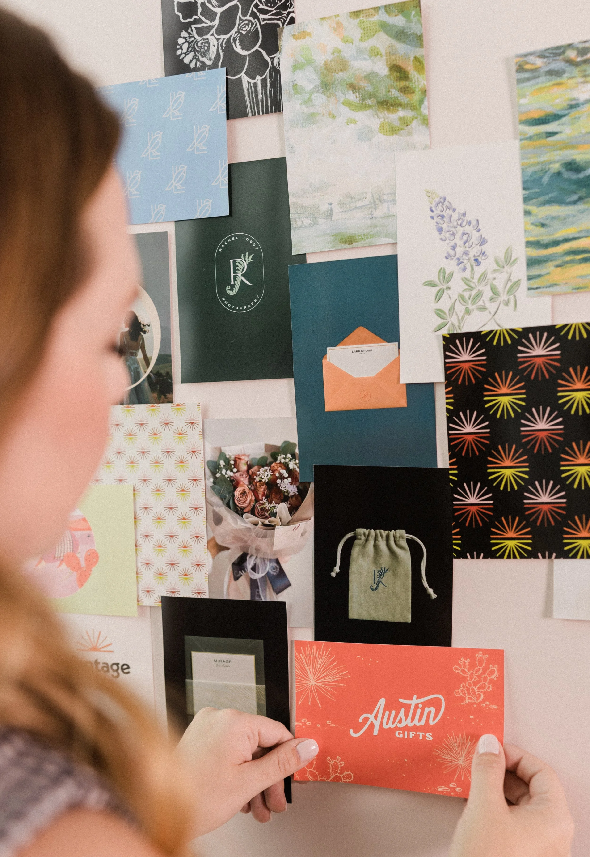 A person holding a red Austin Gifts card in front of a wall decorated with various pieces of colorful paper, cards, and artwork, including floral and abstract designs.