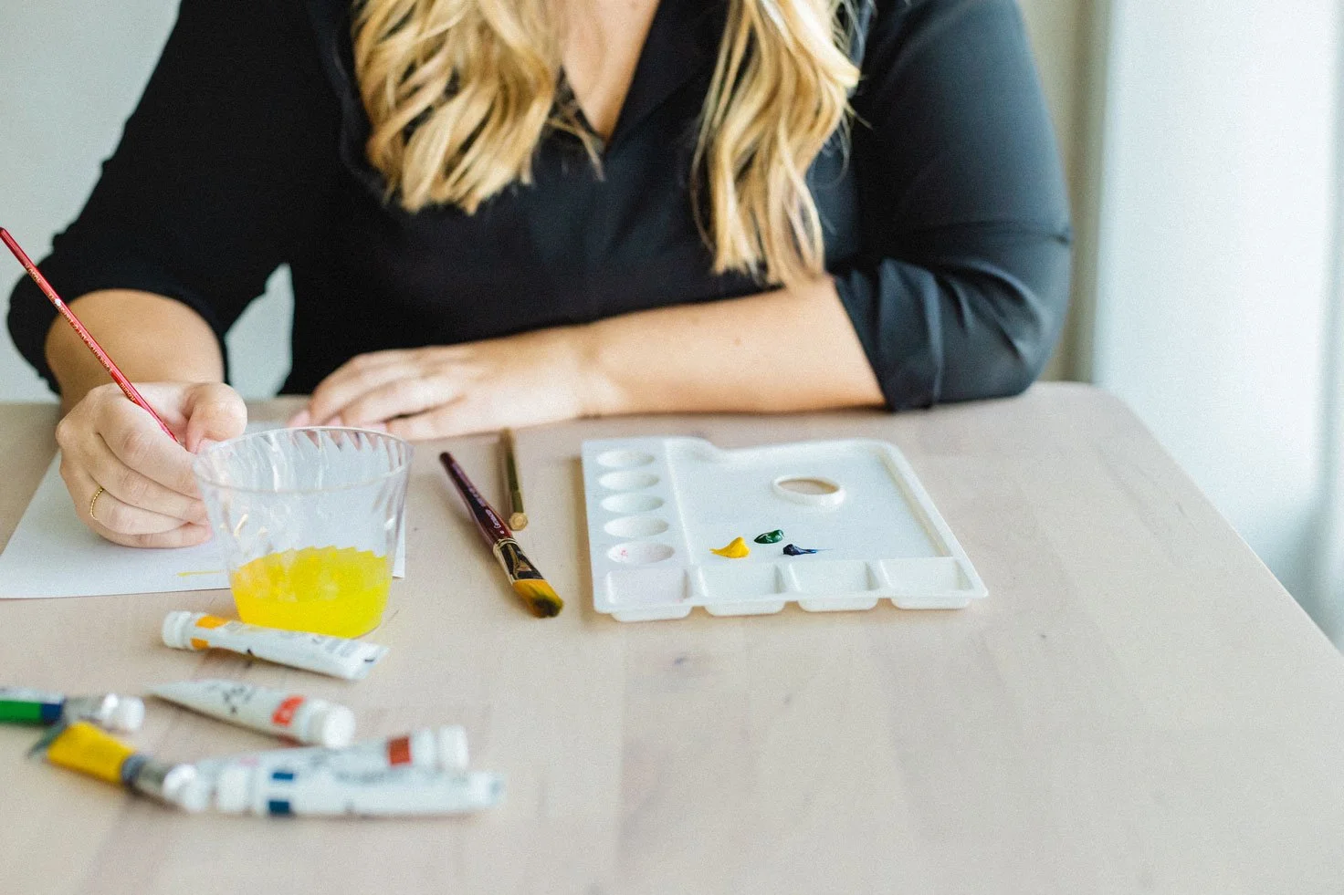 Person with blonde hair in a black shirt paints with yellow and green colors at a table with a paint palette, paints, and a cup of yellow water.