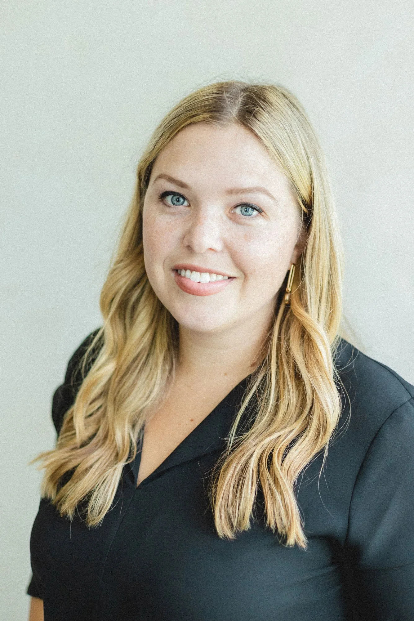 A woman with long blonde hair, blue eyes, and a friendly smile, wearing a black top and gold earrings, standing against a plain light-colored background.