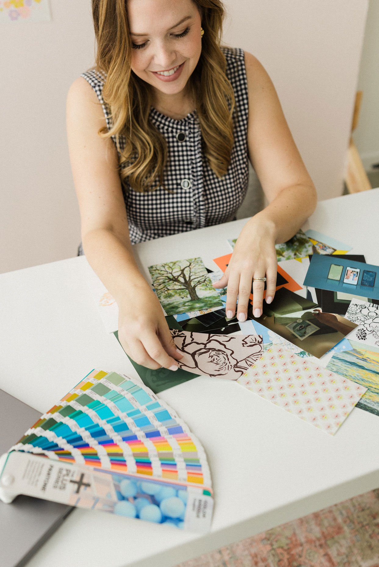 A woman with long, wavy red hair smiling as she sorts through various printed photographs and color swatches on a white table.