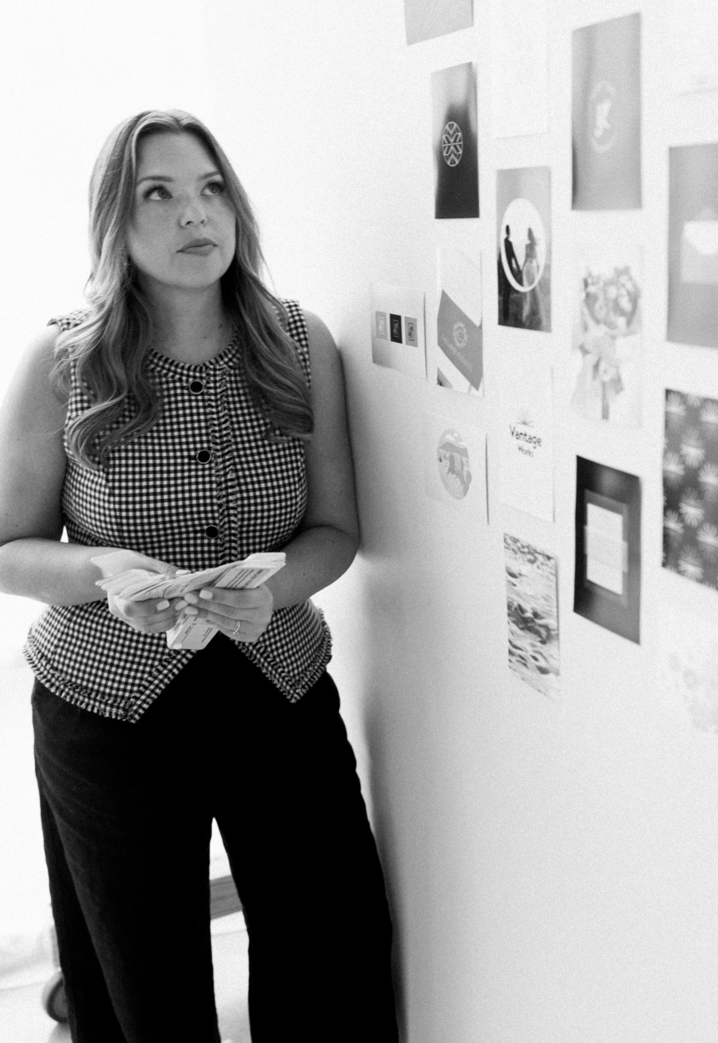 A woman with long hair, wearing a checkered top, stands against a white wall with various images and graphics. She holds some papers in her hands and appears to be observing the wall.
