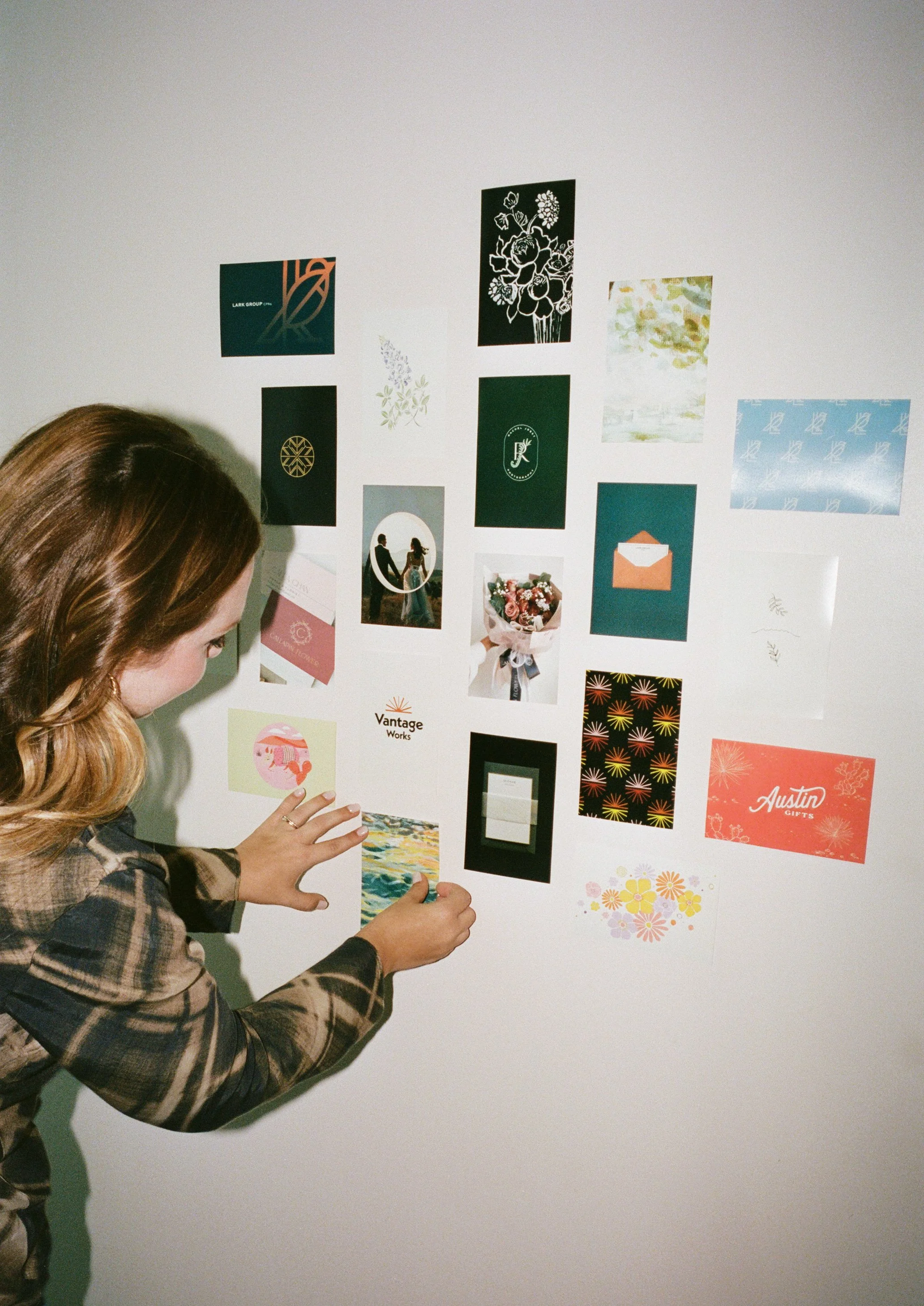 A woman with brown, wavy hair in a plaid shirt is attaching a small colorful print to a white wall among various other images and cards, some featuring floral patterns, illustrations, and text.