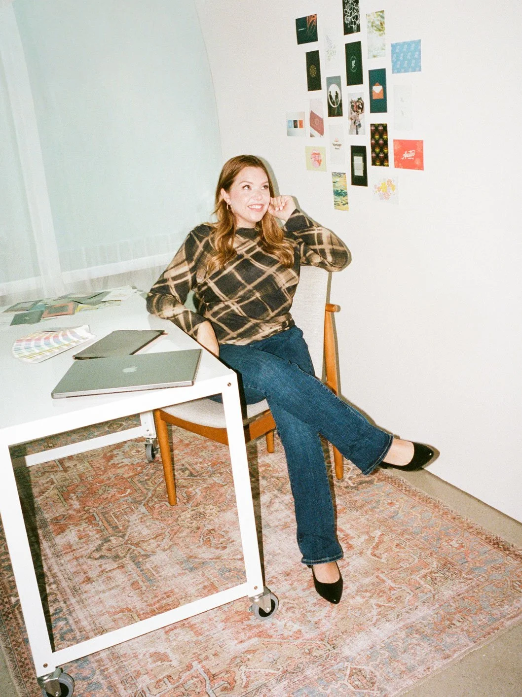 A woman sitting on a wooden chair at a white desk, smiling and posing with one leg crossed over the other. She has long, wavy brown hair and is wearing a black and beige patterned long-sleeved top, dark blue jeans, and black heels. The desk has a closed silver laptop, a black notebook, and color swatches. The background wall has a collage of various colorful photos and prints, and there is a patterned rug on the floor.