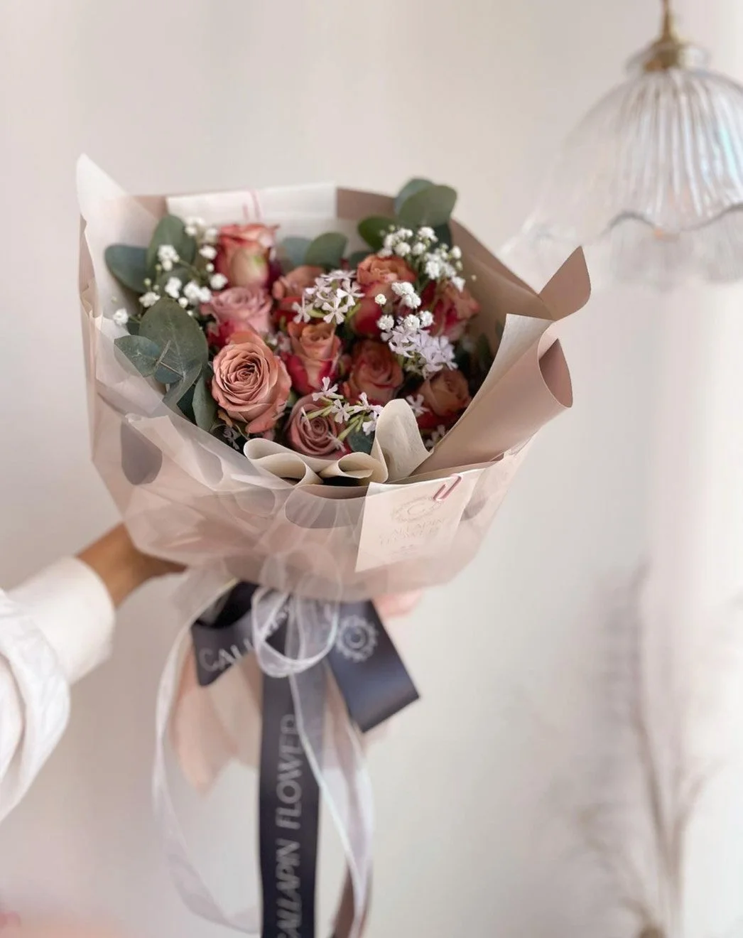 A person is holding a bouquet of pink and peach roses, white baby's breath, and eucalyptus leaves wrapped in pastel-colored paper with ribbon and a tag.