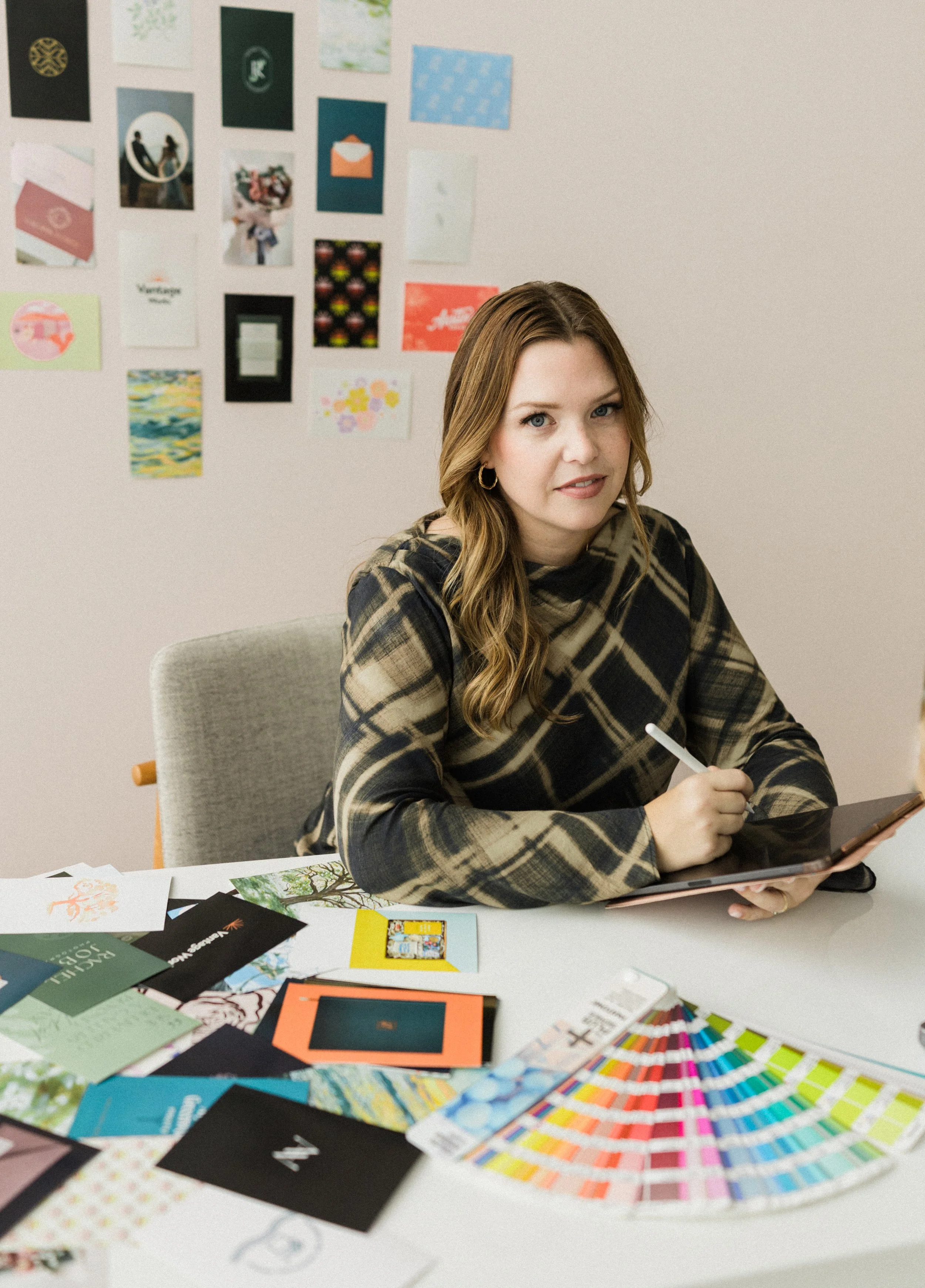 A woman sitting at a table surrounded by color swatches, photo samples, and design materials, holding a tablet and stylus, in a creative workspace with mood boards and cards on the wall behind her.