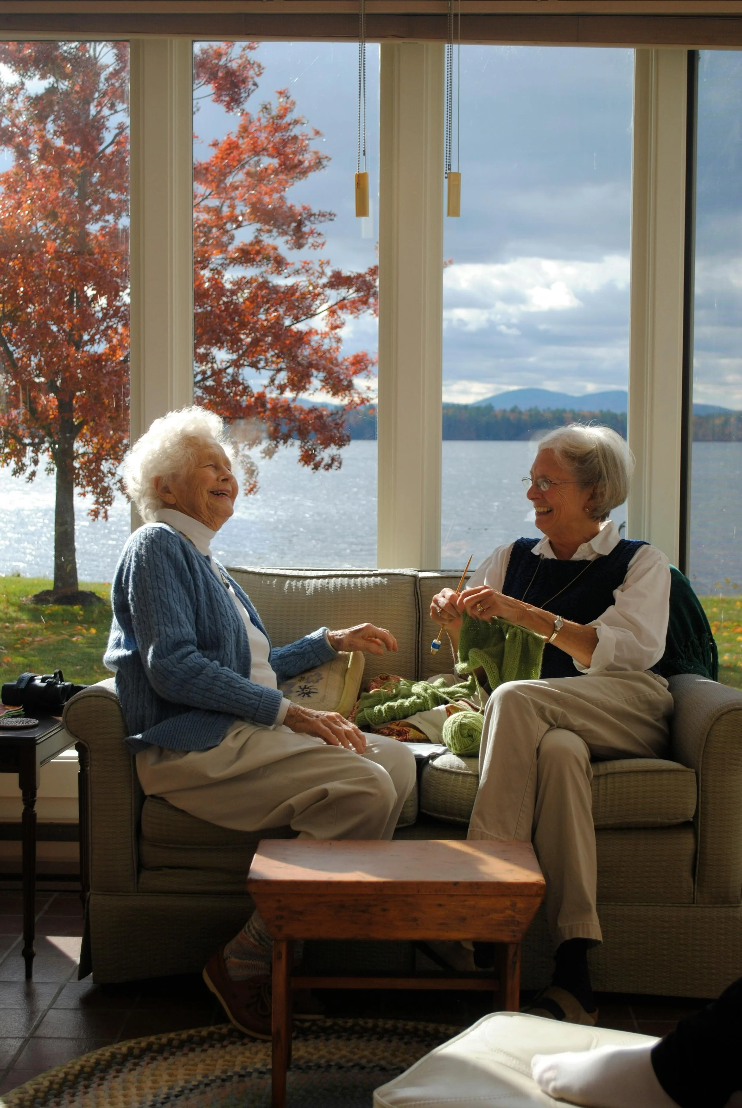 Two elderly women sitting on a sofa by a large window with a lake and autumn trees outside, laughing and knitting.
