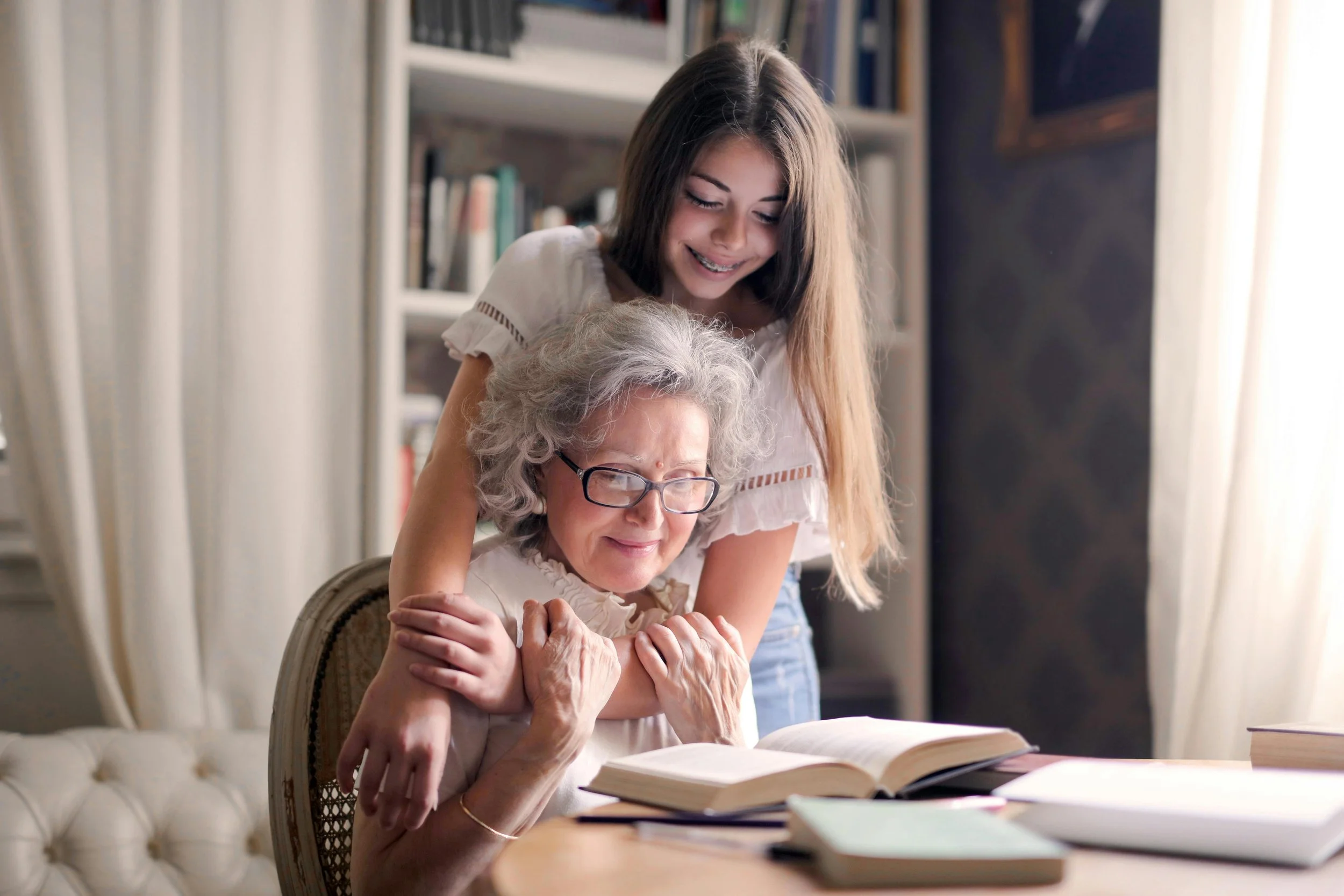 A young woman and an elderly woman are sharing a joyful moment together, reading a book in a cozy, well-lit room with bookshelves in the background.