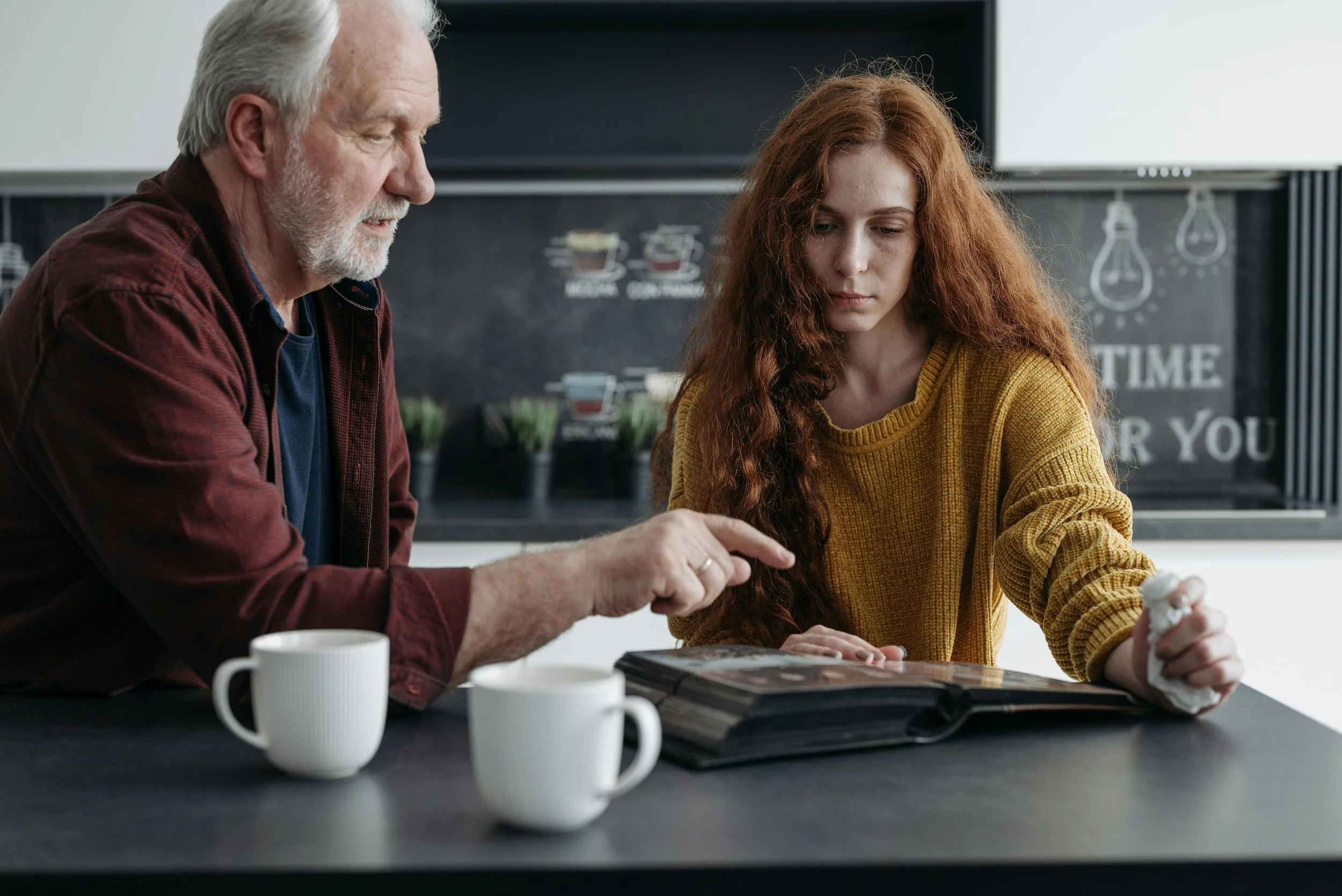 An older man with white hair and a beard, wearing a maroon shirt, is showing a photo album to a young woman with long curly red hair, wearing a mustard yellow sweater, in a kitchen setting with cups on the table.