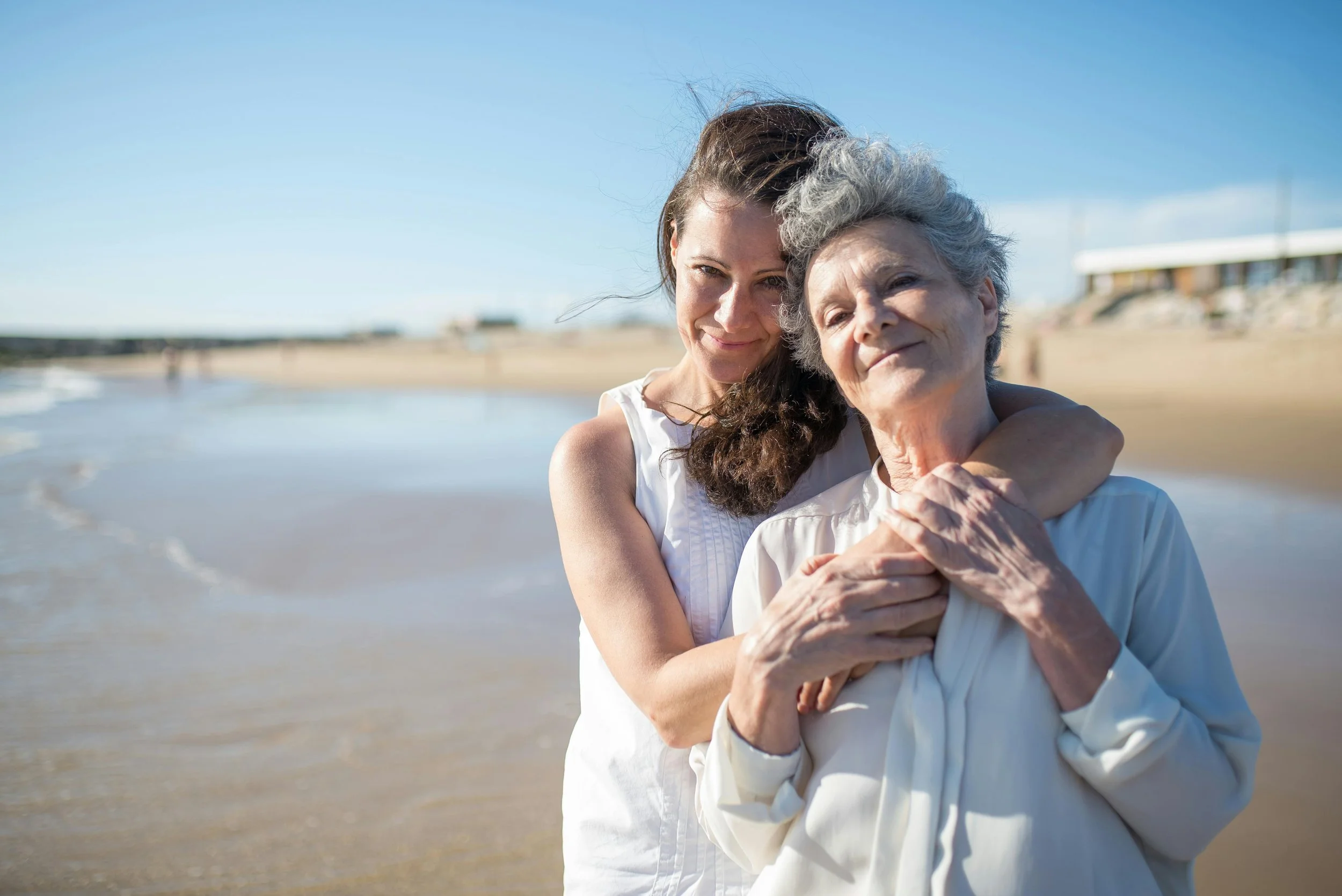 A young woman and an elderly woman embrace on a beach with the ocean and a building in the background.