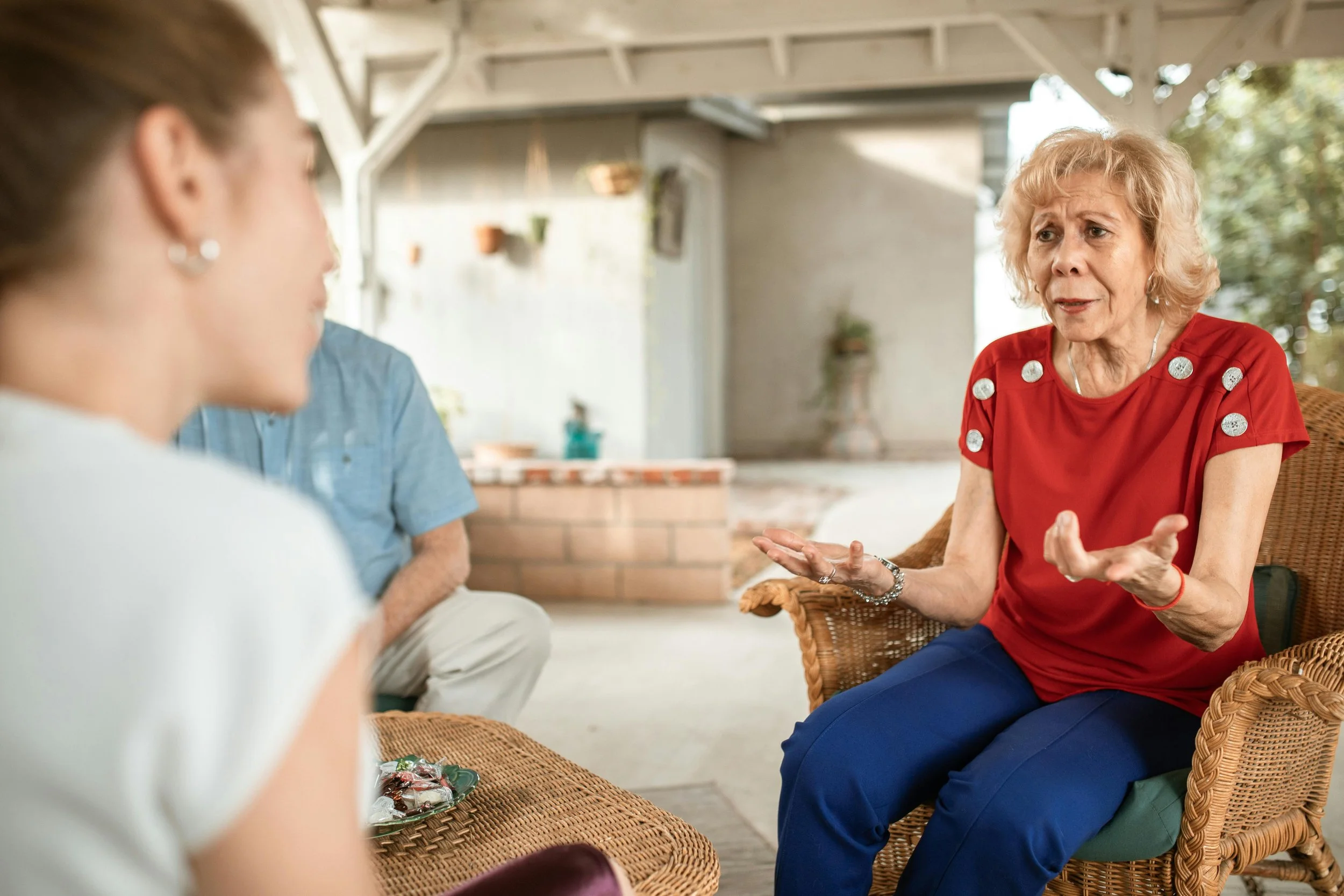 An older woman in a red shirt with decorative buttons on the shoulders is sitting in a wicker chair, gesturing with her hands while talking to two other people who are partially visible. The setting appears to be a covered patio or porch with potted plants and outdoor decor in the background.