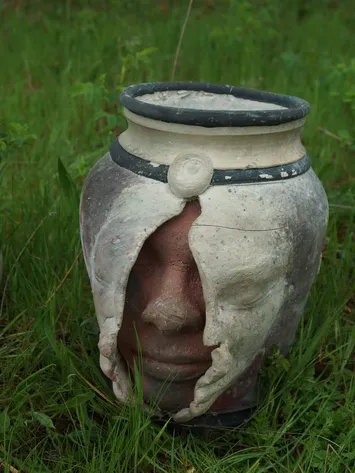 A decorative ceramic vase with a face embedded in it, featuring a large nose, eyes, and lips, placed outdoors on grass.