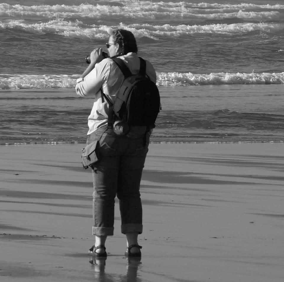 Person standing on the beach, facing the ocean, holding a camera or binoculars, wearing a backpack and sandals.