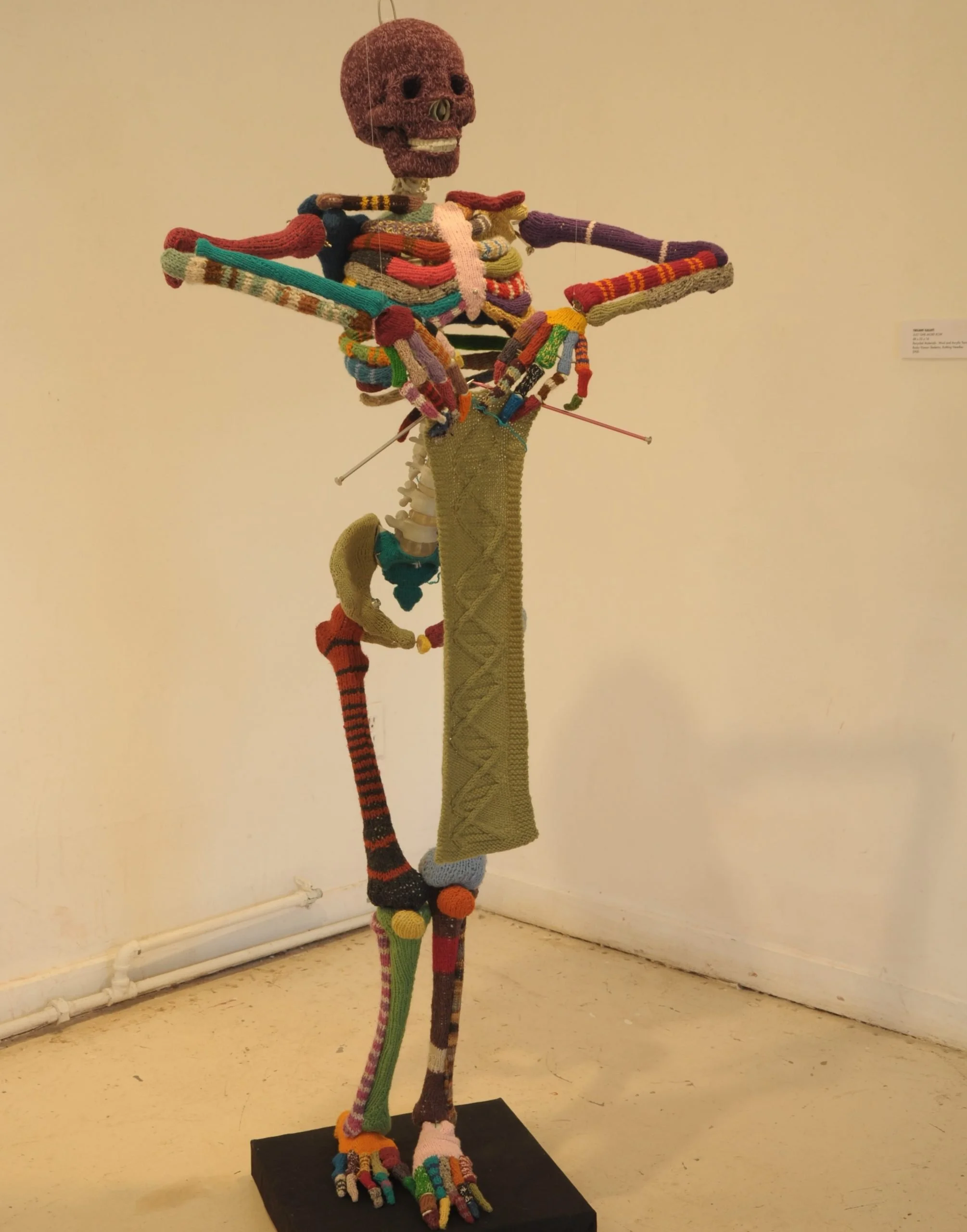 Colorful knitted sculpture of a human figure sitting on a tall pole, with a skull for a head, and with arms outstretched, displayed in a gallery.