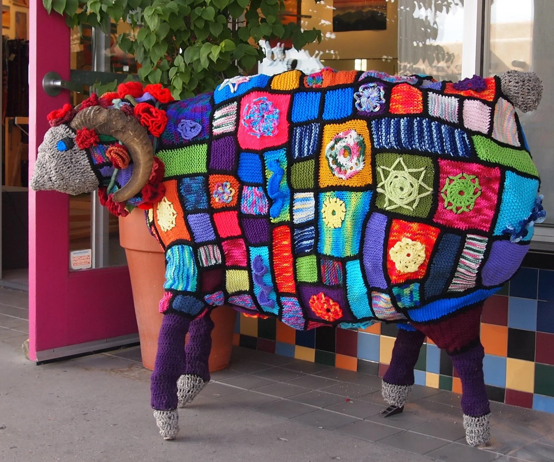 A life-sized sculpture of a cow decorated with colorful crocheted patches and yarn, standing outside a store with a pink door and multicolored tile wall.