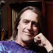 A man with long gray hair and a slight smile, wearing a purple patterned shirt, sitting indoors in front of a wooden bookshelf.