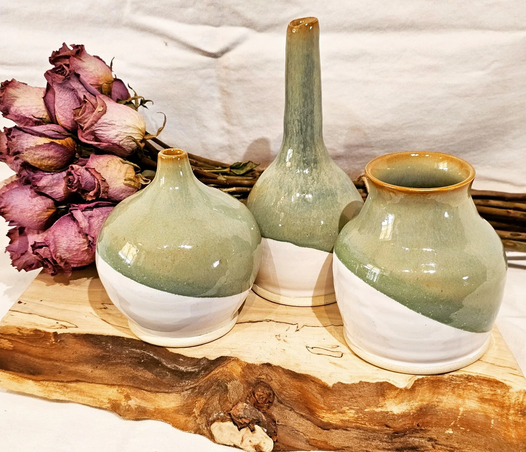 Three ceramic vases with green and white glaze, a bouquet of dried pink roses, and a piece of wooden slab on a light-colored surface.
