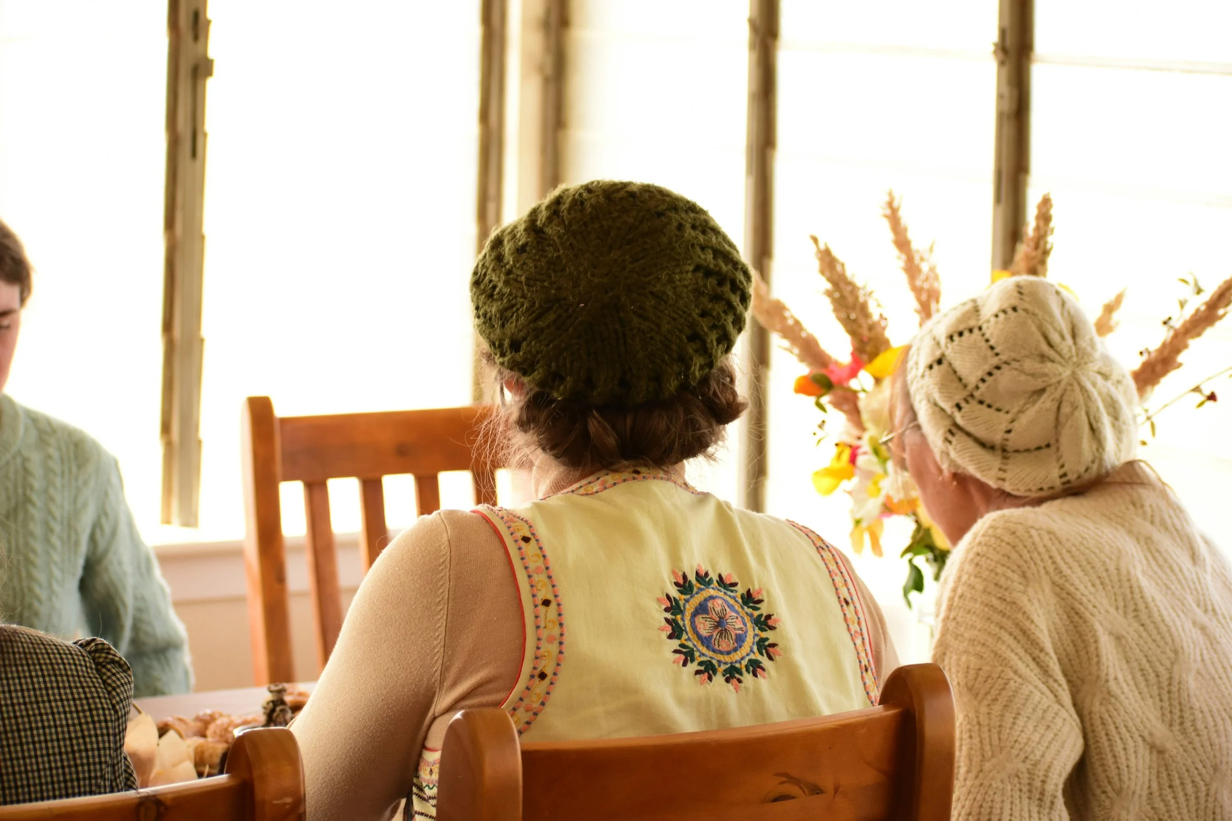 Three women wearing knitted hats sitting at a wooden table with a vase of flowers in a bright room.