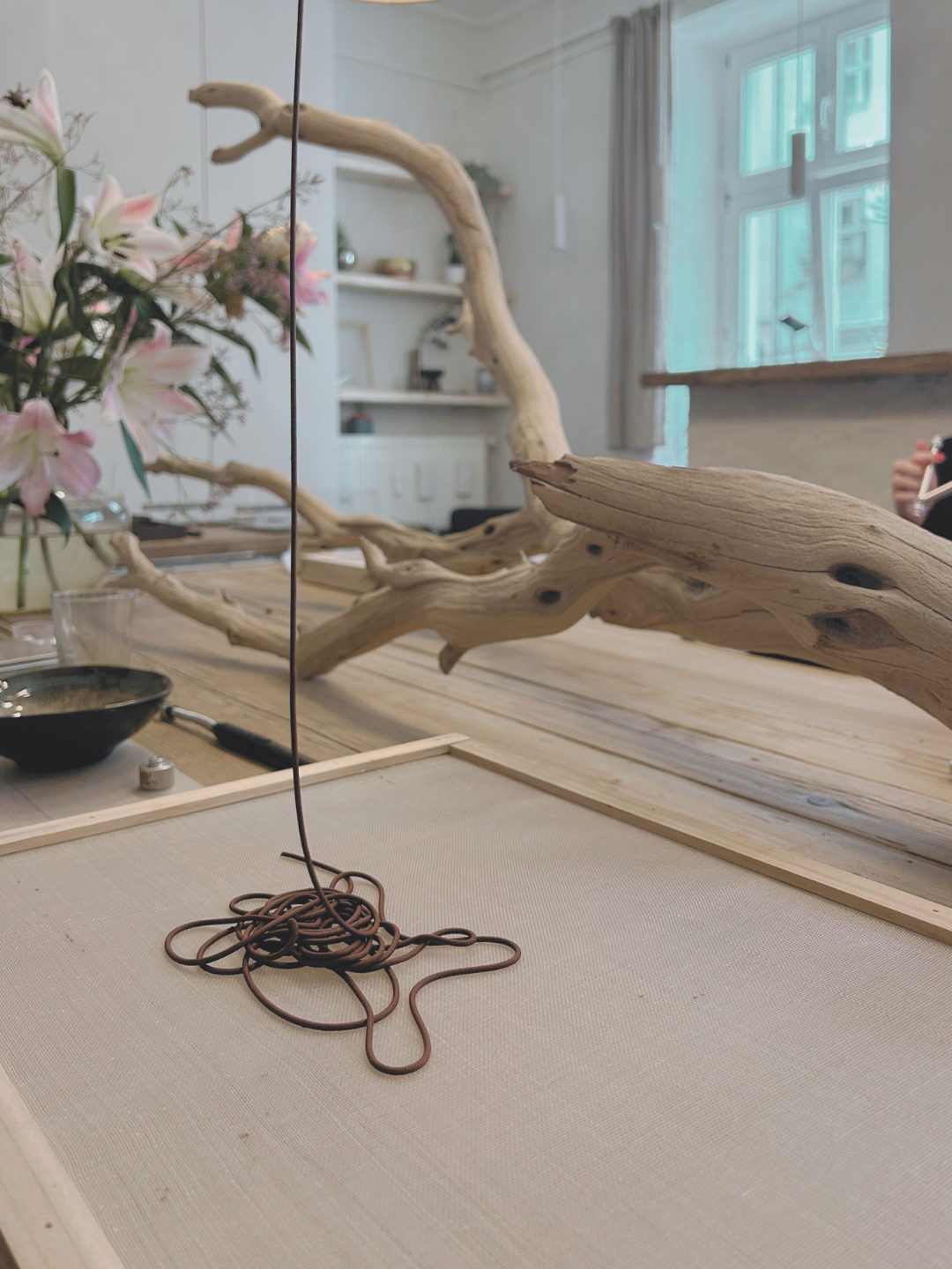 Close-up of a tangled brown cord on a table, with a wood branch and pink flowers in the background in a bright, cozy room with large window.