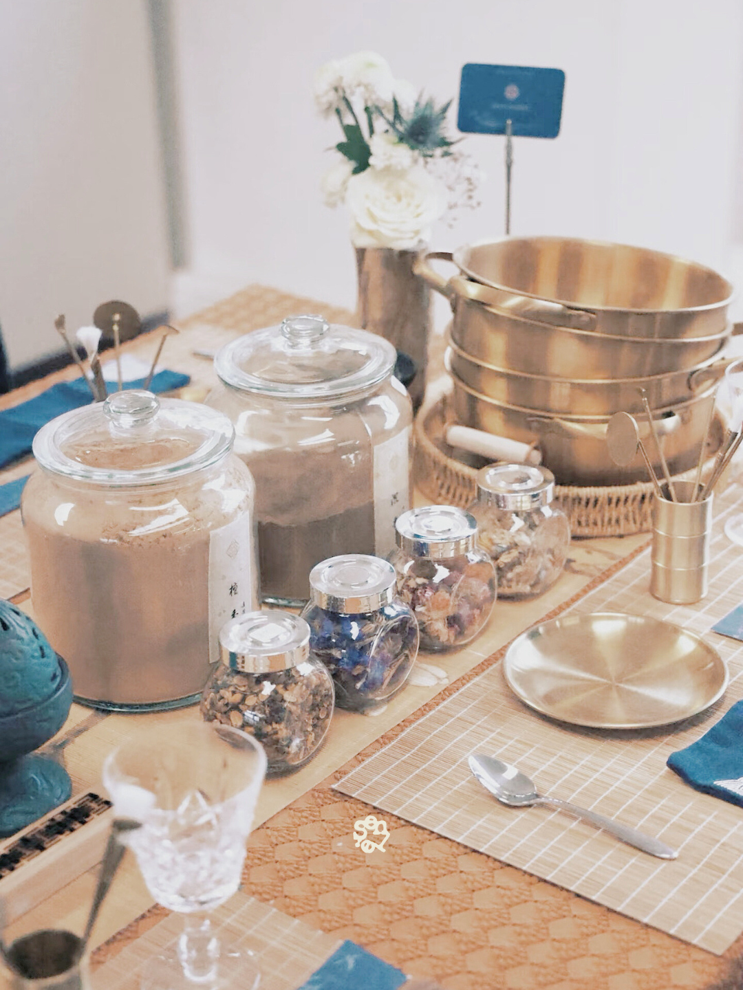 A dining table with glass jars of snacks, brass bowls, a gold plate, a spoon, a small glass, a container with utensils, and a vase of flowers.